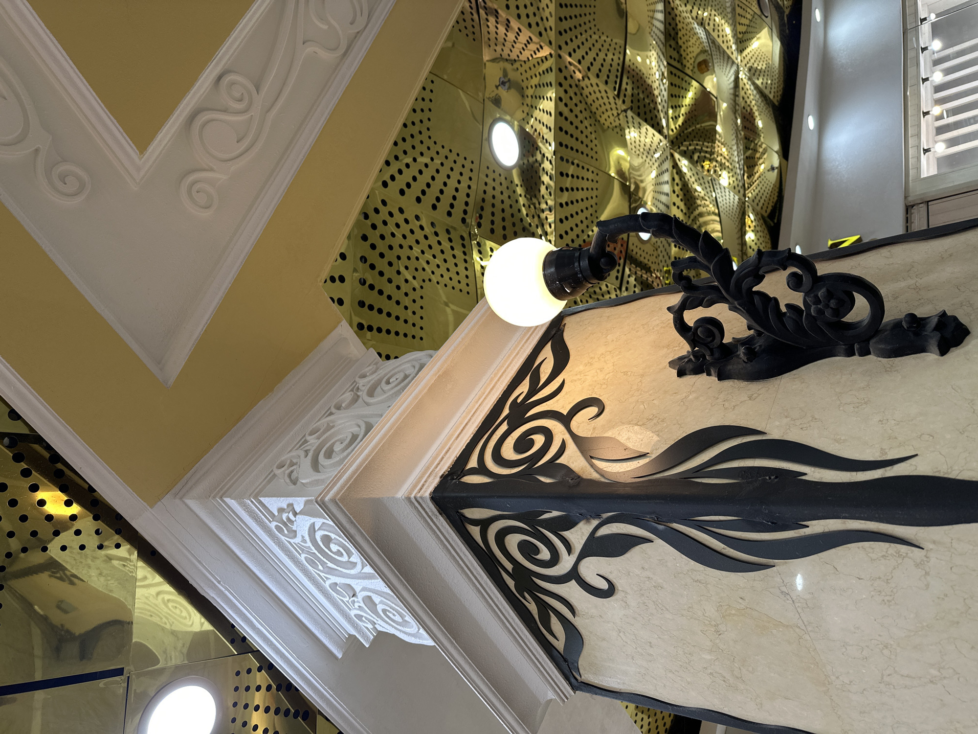A low-angle shot captures architectural and lighting details, possibly from inside a building. The ceiling features ornate plasterwork with scrolled designs, colored yellow and white. Next to it, a reflective surface made of what looks like gold tiles with circular cut-outs forms another part of the ceiling. A black, decorative sconce with a globe light hangs from a beige wall, also ornamented with black, swirling designs. The sconce's light is on, creating a bright spot in the image. The composition includes elements of interior design and architectural detail. Based on the information about the location being the city of Yekaterinburg, Russia, this could be a detailed view of an interior space within a building in that city.