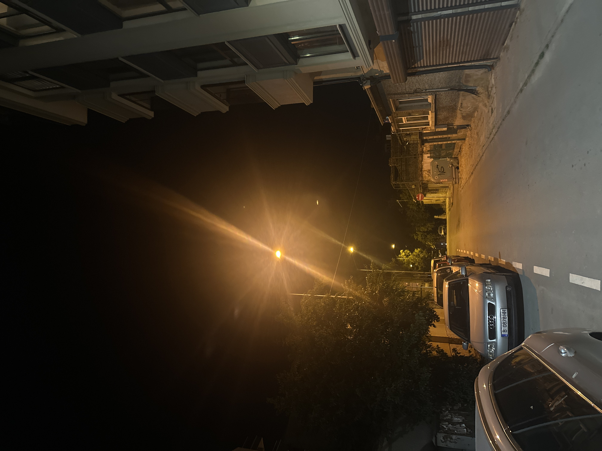 This is a nighttime photograph taken on a street in the city of Varna, Bulgaria. The street is paved and lined with parked cars on the right side. The buildings on the right appear to be commercial or residential, with a large rolling shutter door visible. A tree is present on the left side of the street, casting shadows. Streetlights are illuminated, casting a warm glow and creating lens flares in the image. The sky is dark, indicating it is nighttime. One of the parked cars, a silver Audi, has a license plate with the characters "B 0878 HH".