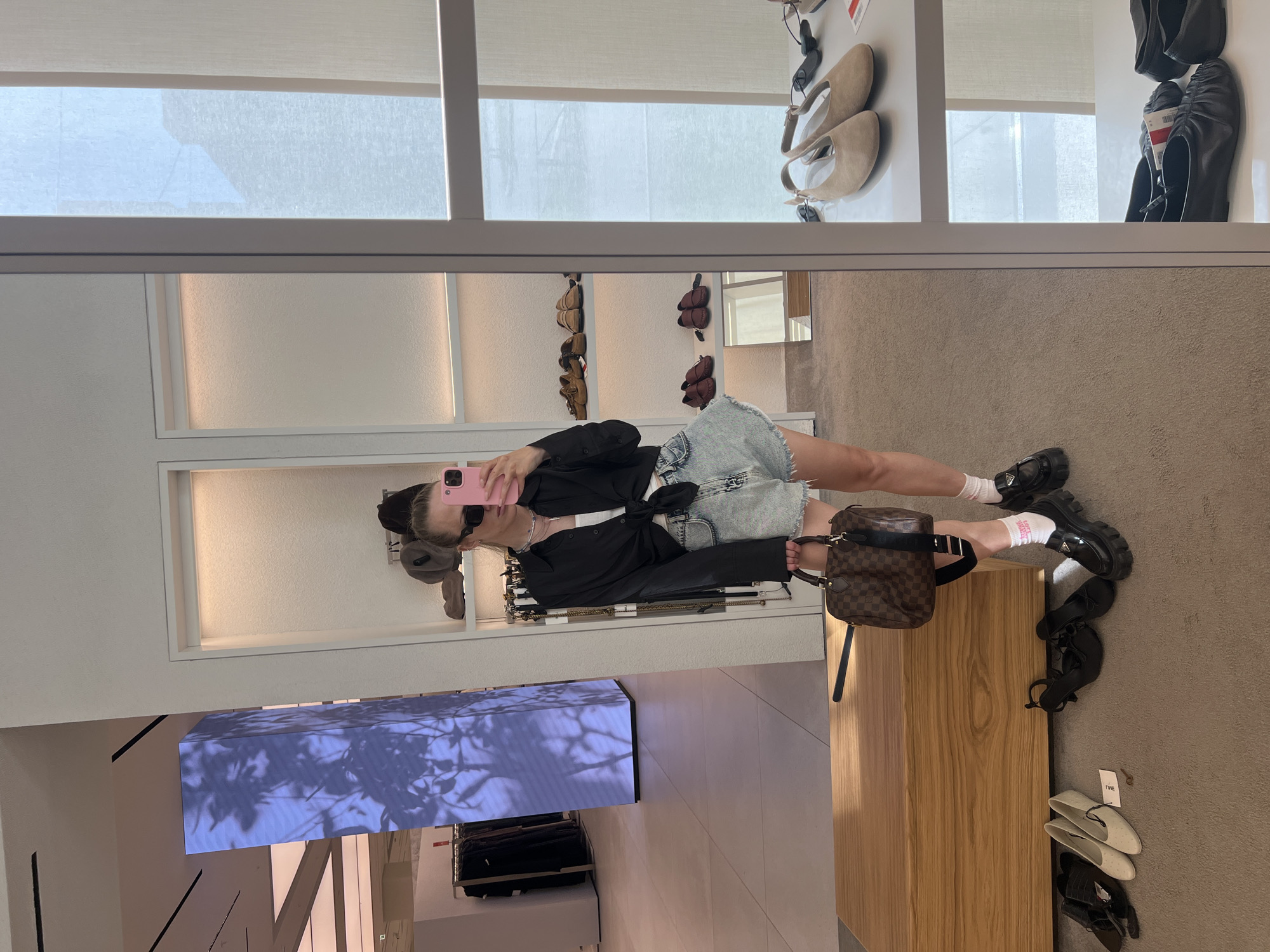 A young woman stands in a retail store, taking a mirror selfie. She is wearing a black collared shirt tied at the waist, a white tank top, denim shorts, white socks, and black chunky loafers. She carries a brown checkered designer handbag. Her hair is pulled back, and she is wearing sunglasses and holding a pink smartphone. The store appears to be a clothing and shoe boutique, with displays of clothing and shoes visible in the background and in a large mirror reflecting the scene. The lighting is bright and artificial. The floor is carpeted. The overall impression is that of a fashion-conscious individual shopping in a modern retail environment. The location of this store in Saint Petersburg, Russia cannot be determined from the image.
