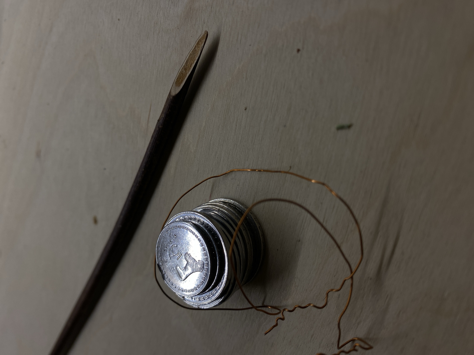 The image depicts a stack of approximately 15 Georgian 5 Tetri coins, a length of thin, bare copper wire, and a dark brown, sharpened wooden stick, all lying on a light-colored, seemingly wooden surface. The coin displays a griffin. The scene appears to be indoors under diffuse lighting. No information to suggest the location is Tbilisi, Georgia is present in the image.