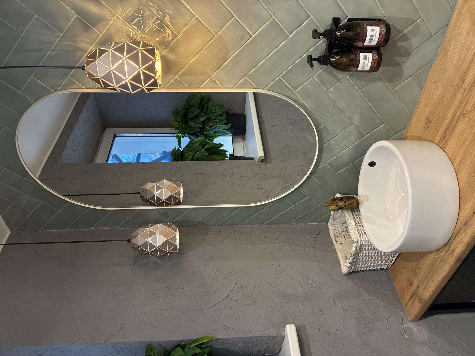 This is a medium shot of a bathroom sink area with a rustic and modern aesthetic, set in Unknown, Unknown. The dominant wall to the right of the scene is covered in a herringbone pattern of glossy, pale green tiles. To the left, a wall has a textured grey finish.
The sink is a white, round vessel sink with a matte gold faucet. It sits on a light-colored wooden countertop with a visible grain. To the left of the sink is a small, white woven basket with a floral fabric lining.
Above the sink, an oval mirror with a thin, white frame is hung. The mirror reflects two black pendant lights with geometric, triangular metal shades, as well as two potted green plants on a windowsill visible in the reflection. The pendant lights are suspended from the ceiling.
On the tiled wall to the right, a black dispenser rack holds two dark brown pump bottles with white labels that appear to be for hand soap or lotion. The bottles have ornate, patterned labels.
The lighting is warm and casts subtle shadows, highlighting the textures of the walls and the sheen of the tiles. The overall impression is one of a stylish and functional bathroom space.