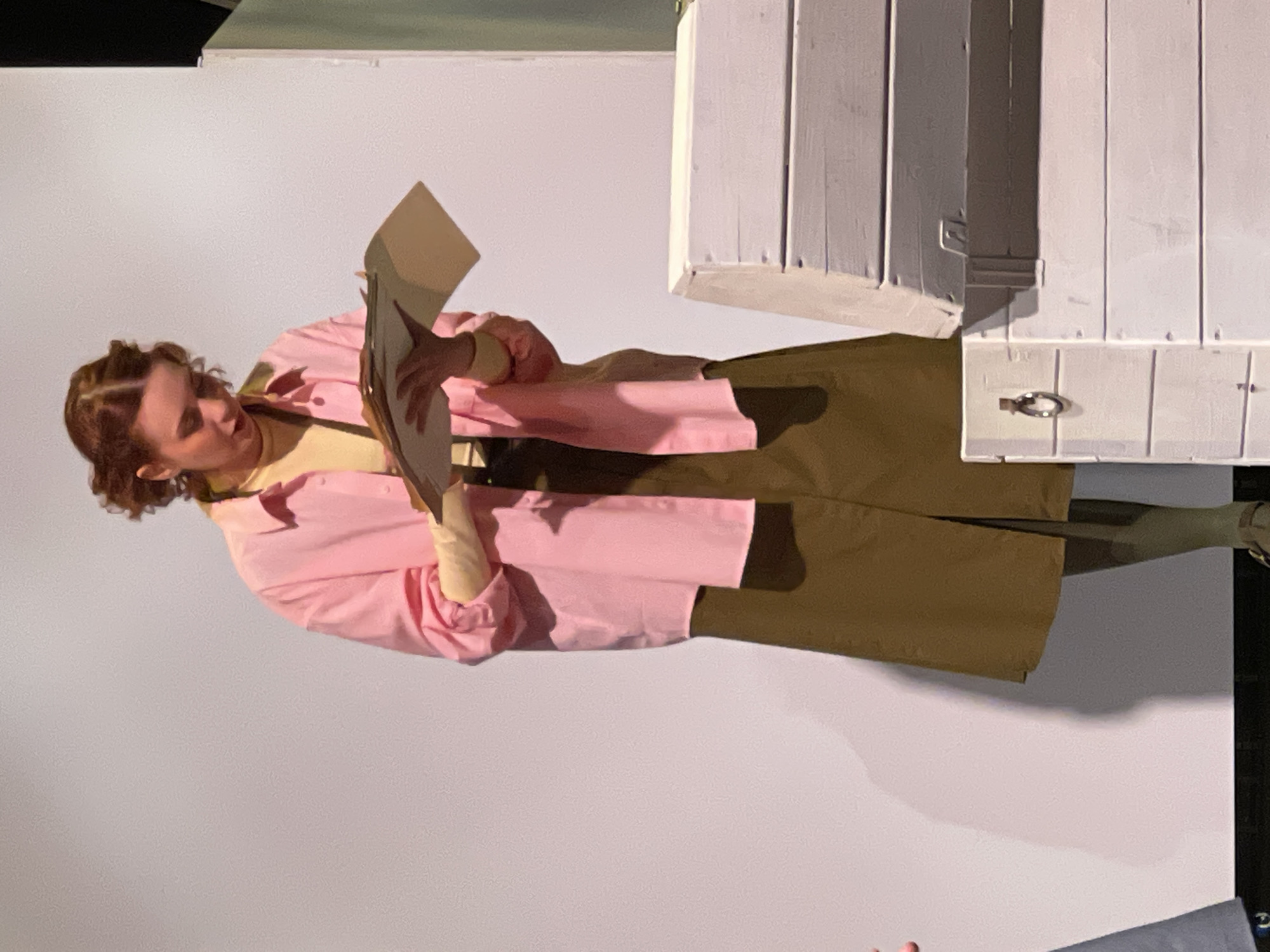 A woman stands reading a document near a white wall and a partially visible white wooden structure with a small metal handle. She wears a pink jacket over a yellow top and a khaki-colored skirt. The image appears to be taken indoors. The city of Kaliningrad, Russia, is not visibly identifiable from the provided image.
