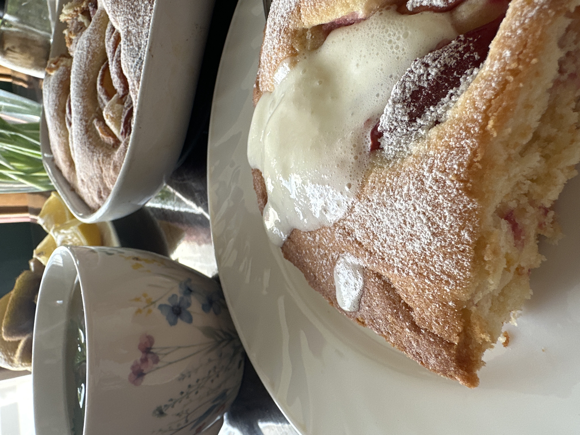 The image shows a piece of cake on a white plate, topped with a creamy white sauce and sprinkled with powdered sugar. Next to the plate is a cup with a floral design, possibly containing tea or coffee. In the background, another cake is visible in a white baking dish. The scene appears to be set indoors, possibly at a table. There is no specific information to suggest the location is in Chaikovsky, Russia, or any details about people, interactions, or environment beyond what is visible.