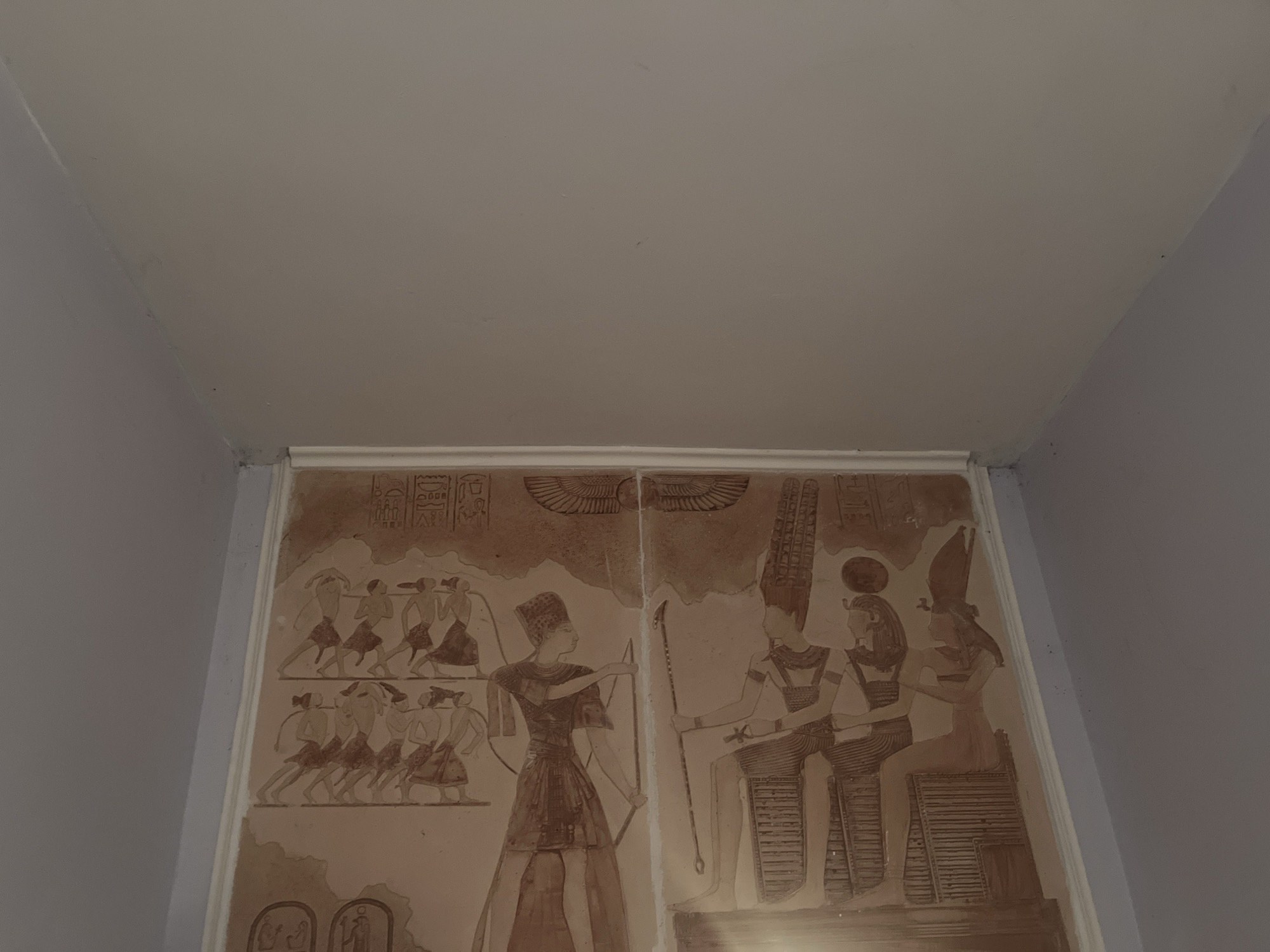 The image shows a wall panel with Egyptian-style artwork. The panel depicts human figures and hieroglyphs. One section shows a figure with a bow and arrow aiming at a group of people running. Another section features seated figures wearing royal attire. The panel appears to be installed in a room, as indicated by the adjacent walls and ceiling. It is unlikely the location is Goce Delchev, Bulgaria, given the style of artwork.