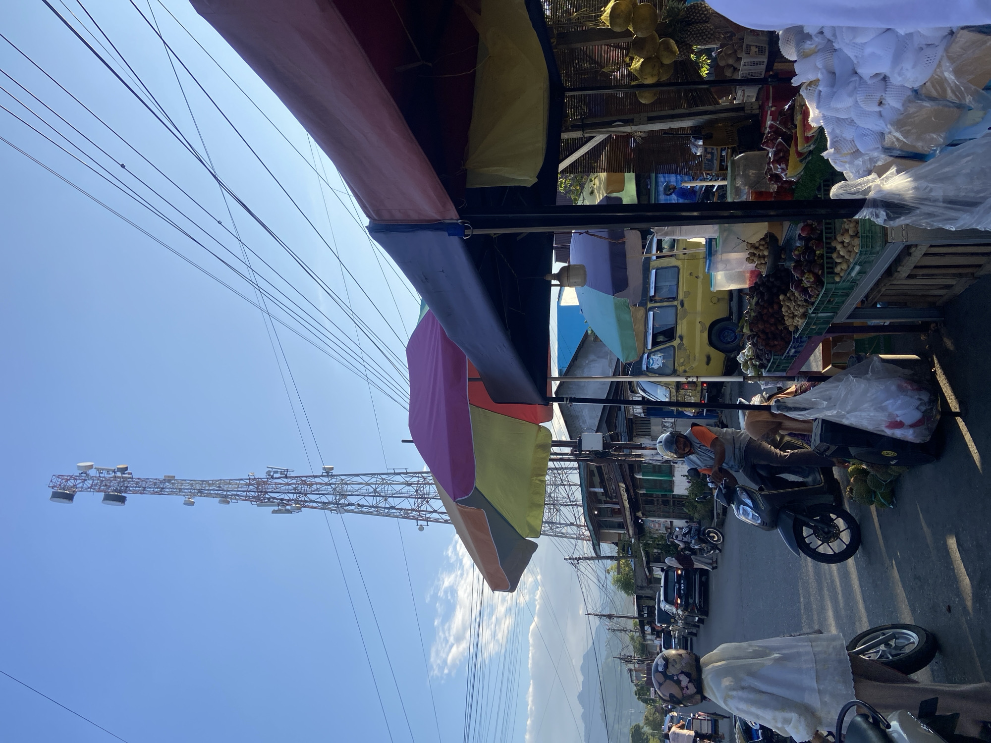 The image depicts a street scene in Palopo, Indonesia, likely during the daytime. A tall telecommunications tower is prominently visible in the background against a clear blue sky. In the foreground, a roadside market is set up under colorful awnings. Various fruits, including rambutans, mangosteens, and pineapples, are displayed for sale. Several people are present, some riding motorcycles and others browsing the market stalls. Cars and motorcycles are also seen on the street, suggesting a moderately busy urban environment. The architecture visible in the background appears to be of a tropical, possibly Southeast Asian, style.