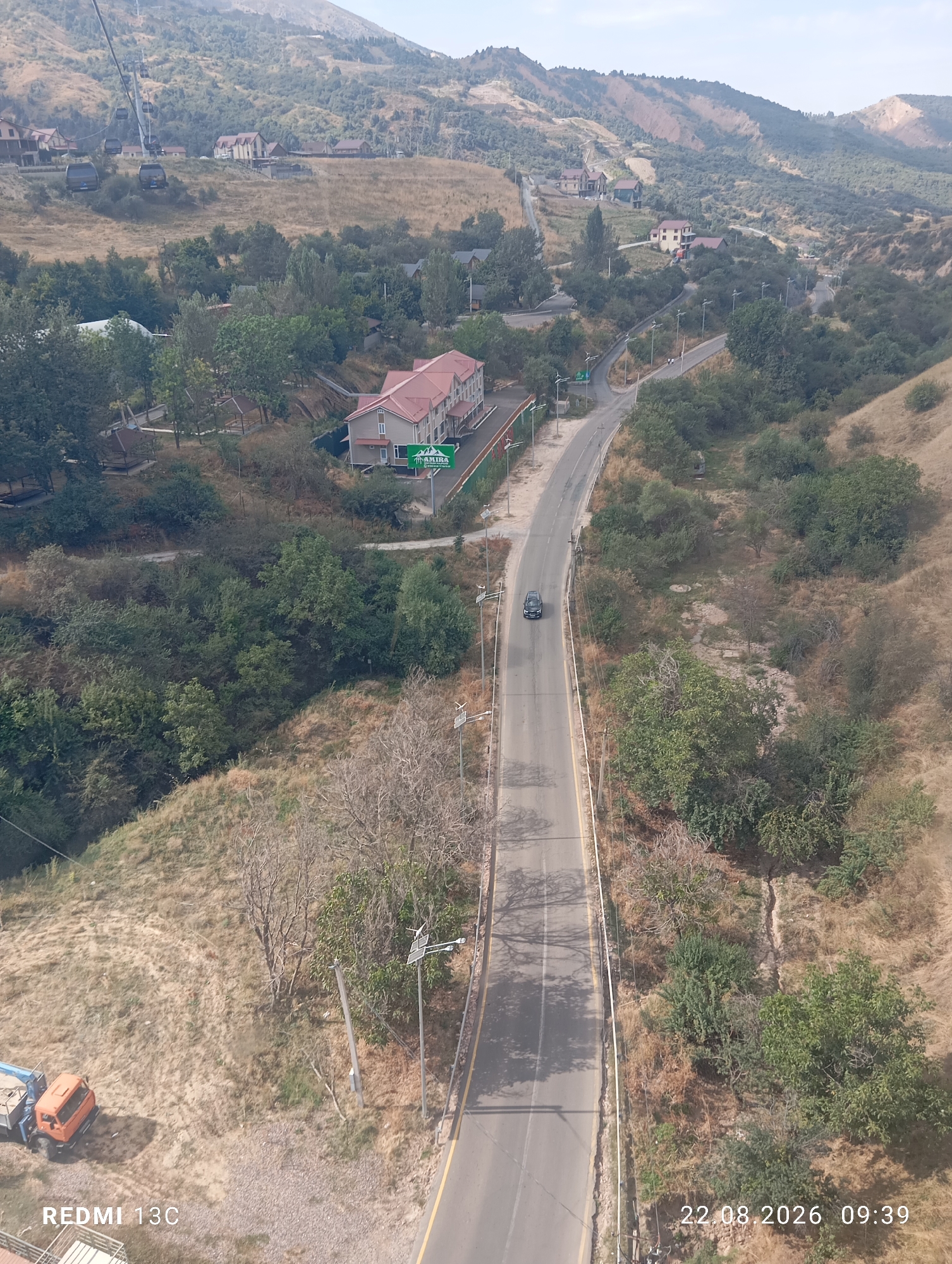 The image is a high-angle aerial view of a mountainous area in Amirsay, Uzbekistan, likely taken from a cable car. A paved road winds through the scene, with a single black car driving on it. The landscape is characterized by dry, grassy hillsides dotted with various trees and shrubs. In the middle ground, a multi-story building with a red roof is visible, along with several smaller structures and gazebos. Further up the hillside, more buildings and a ski lift system can be seen. The sky is overcast. In the lower left corner, a small orange truck is parked near some vegetation. Timestamp information in the bottom right corner indicates the image was taken on August 22, 2026, at 09:39. The text "REDMI 13C" is also present in the lower left corner, likely indicating the device used to capture the image.