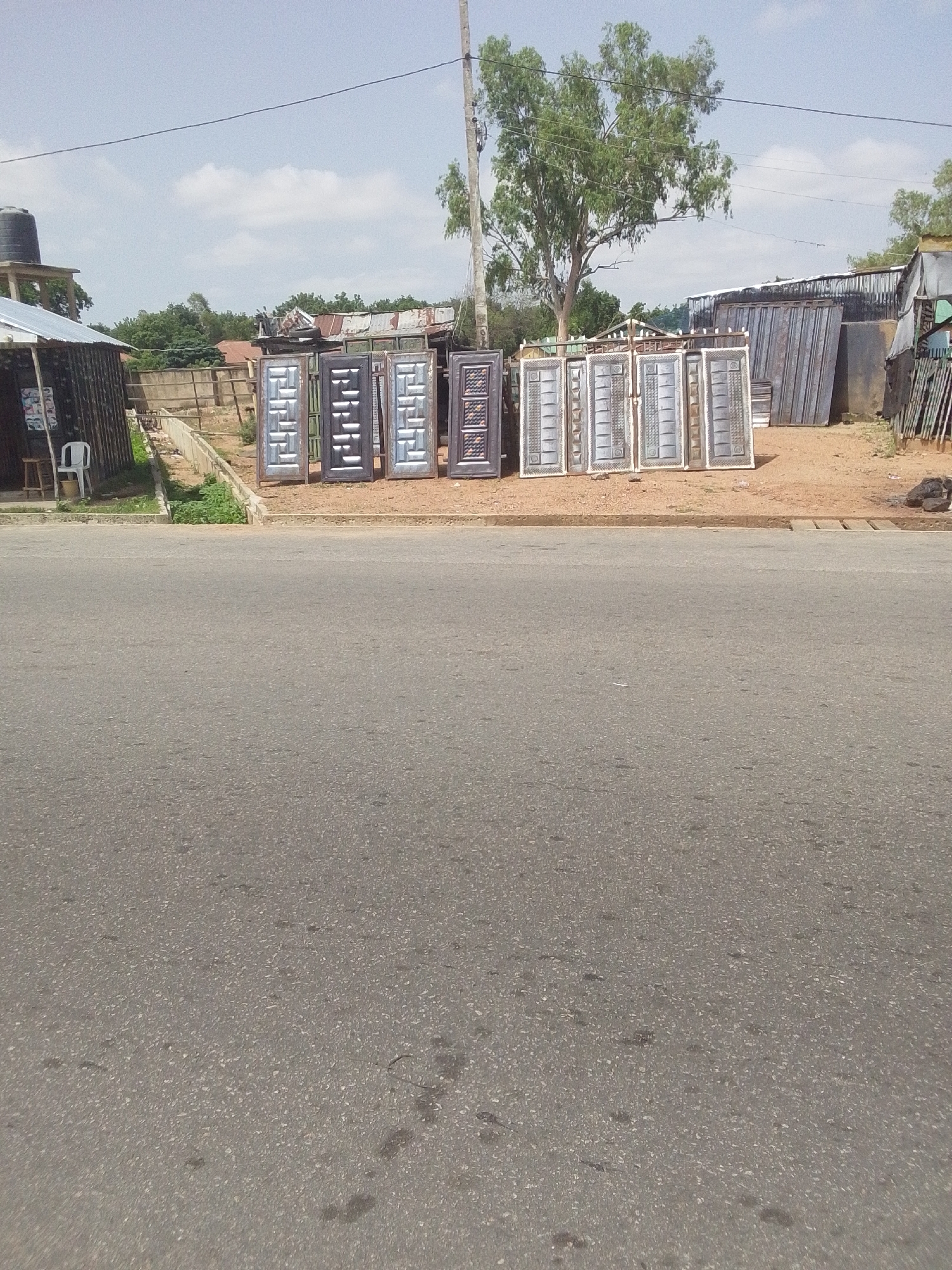 In Jalingo, Nigeria, along a street, a collection of metal doors is displayed outside a small business. The doors, featuring decorative patterns, are arranged in a line on a dirt surface. Behind the doors, a modest structure with a corrugated metal roof is visible, along with some stacked metal sheets and other building materials. A large tree with green foliage stands behind the business, under a partly cloudy sky. The foreground of the image is dominated by a paved road.