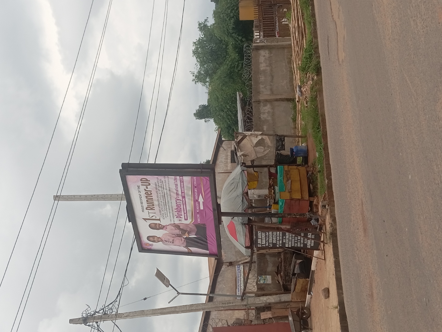 This image, taken in Nkpor, Nigeria, captures a daytime street scene under an overcast sky. The primary subjects are a large billboard and a roadside stall, positioned alongside a paved road with a dirt shoulder.
The billboard, centrally located, advertises "Pinkberry International School." It proudly proclaims the school as the "PROUD RECIPIENT OF THE 1ST Runner-UP" in the "BEST APPROVED PRIVATE PRIMARY SCHOOL CATEGORY" and also mentions being "1ST RUNNER UP IN BEST TEACHERS AWARD...IN NIGERIA 2025." The billboard features an image of two young girls in pink school uniforms. Contact details provided include "OBOSI ANAMBRA STATE TEL:0803887120" and the services offered: "CRECHE NURSERY PRIMARY COLLEGE."
To the right of the billboard, a multi-purpose roadside stall is visible. It has a makeshift structure with a white tarp covering part of it, and various colored containers or boxes are stacked around. A sign attached to this stall reads "ST JOHN AGENCY & MANAGEMENT," followed by "Always Trust in God" (Marts Trust in God per OCR) and phone numbers: "08063846494" and "09049725413." The sign also advertises services such as "ROOMS TO RENT," "SELF CONTAINED EXTENSION," and "AMBULANCE."
In the background, a concrete fence topped with barbed wire runs parallel to the road, with lush green trees visible beyond it. Overhead, numerous power lines crisscross the sky, supported by utility poles. The street itself appears to be a thoroughfare, suggesting an active urban or semi-urban environment. The cloudy sky indicates a likely overcast or non-sunny day.