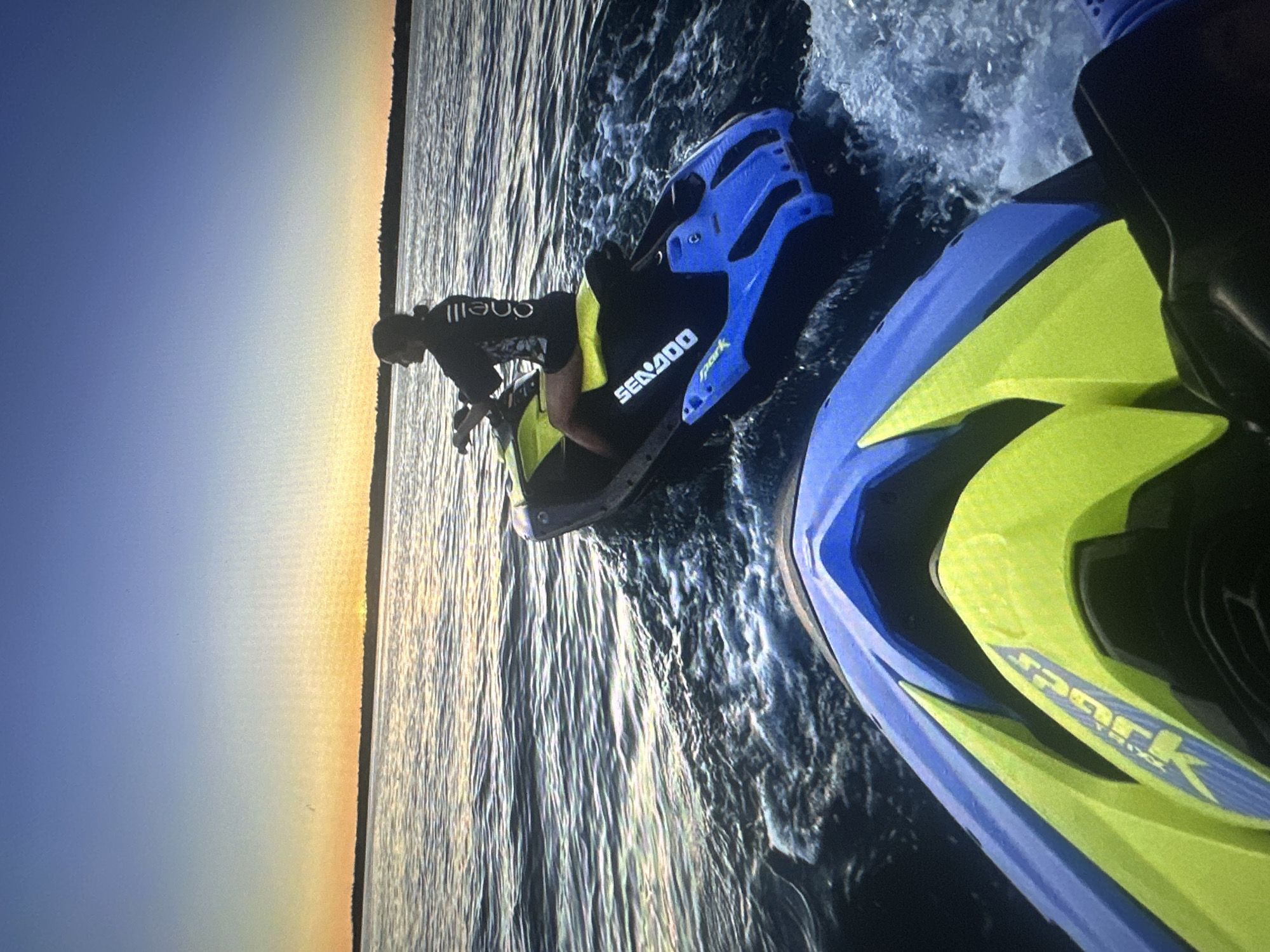 Two jet skis are navigating on the water during sunset. The jet ski in the foreground is a Sea-Doo Spark, predominantly blue and neon yellow. The jet ski in the background is also a Sea-Doo Spark, black, blue, and neon yellow, with a rider. The rider is wearing a black O'Neill wetsuit top and dark shorts. The jet skis are creating wakes as they move through the water. The sky is a gradient of blue to orange, indicating a sunset. The horizon shows a dark line of trees or land. Based on the distinct sunset coloration and the prevalence of clear water activities common in this region, this scene is likely taking place on the Baltic Sea, possibly near the coast of Tallinn, Estonia.