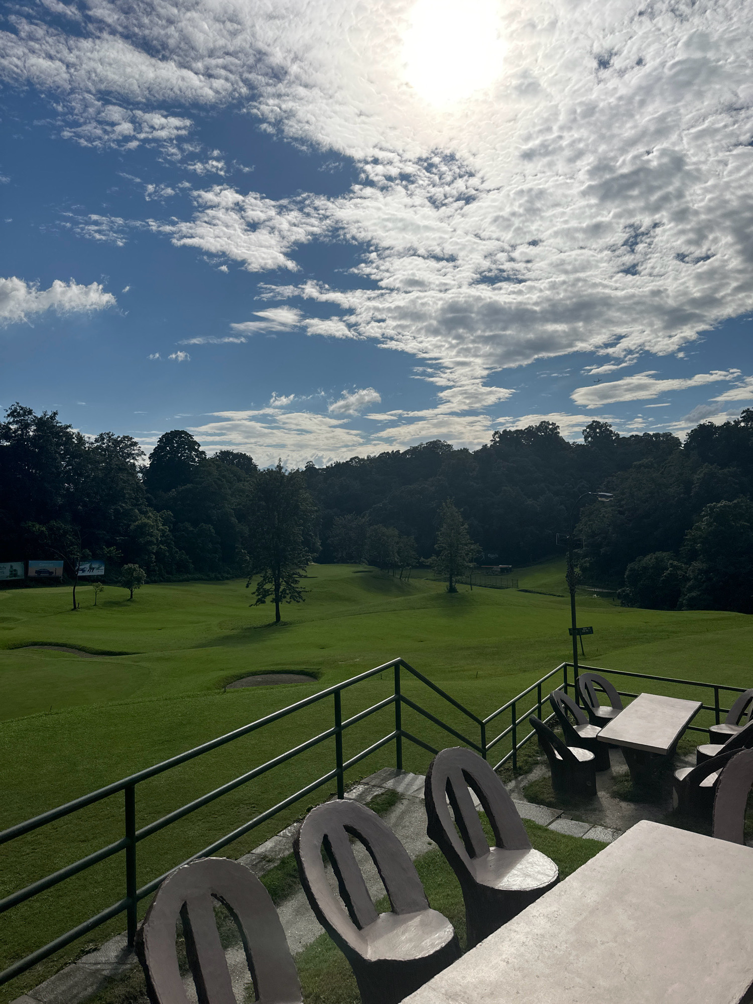 The image shows an outdoor seating area overlooking a golf course in Kageshwori Manohara, Nepal. In the foreground, there are concrete tables and chairs arranged on a patio. A metal railing separates the patio from the golf course. The golf course itself is lush green with sand traps and trees. In the background, a dense forest covers rolling hills. The sky is blue with scattered white clouds, and the sun is visible, casting a bright light. The overall scene depicts a serene and picturesque setting, likely a restaurant or clubhouse with a view of the golf course.