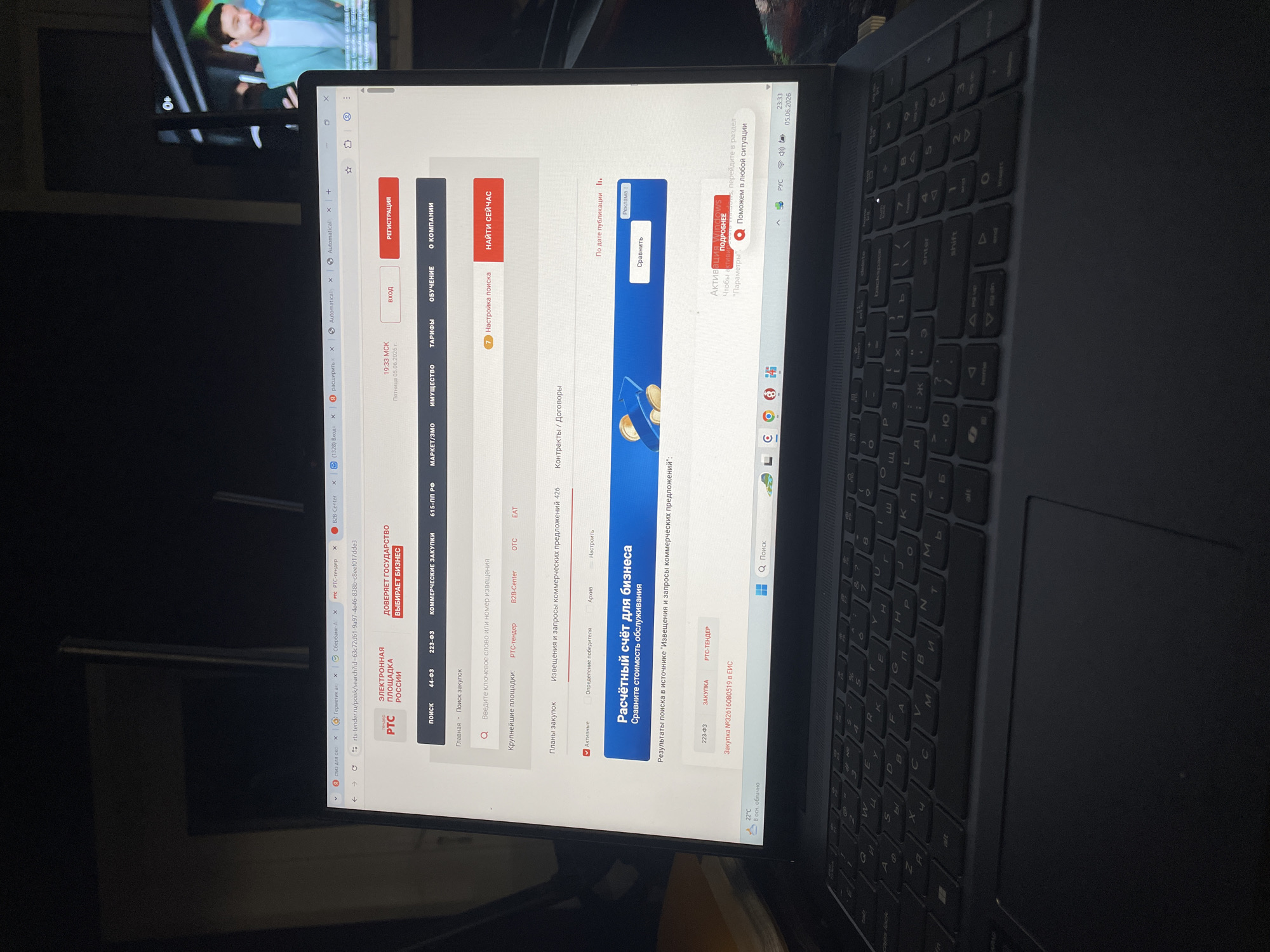 A forensic examination of the image reveals an indoor scene in Chulym, Russia, depicting an open, black HP laptop positioned on a dark surface, likely a desk or table. The laptop screen displays a web browser (Microsoft Edge) actively viewing the Russian procurement website rts-tender.ru, specifically a search results page for "Закупка №32616080519 в ЕИС" (Procurement №32616080519 in EIS). The website's displayed time is "19:33 МСК" (Moscow Standard Time) on "Пятница 05.06.2026 г." (Friday 05.06.2026). The laptop's taskbar shows the system time as "23:33 05.06.2026," indicating a 4-hour difference from Moscow Standard Time, consistent with the UTC+7 time zone of Chulym, Russia. The taskbar also displays a weather icon showing "22°C В осн. облачно" (mostly cloudy) and a "РУС" (Russian) language setting. A "Активация Windows" (Windows Activation) pop-up is partially visible on the screen. The laptop features a black QWERTY keyboard with Cyrillic characters and a numeric keypad. In the background, to the right of the laptop, a flat-screen television is displaying a male figure with dark hair, wearing a light blue jacket over a white shirt, smiling and looking left. A "0+" age rating logo is visible in the top right corner of the television screen. The room appears dimly lit. No individuals are present within the immediate scene.