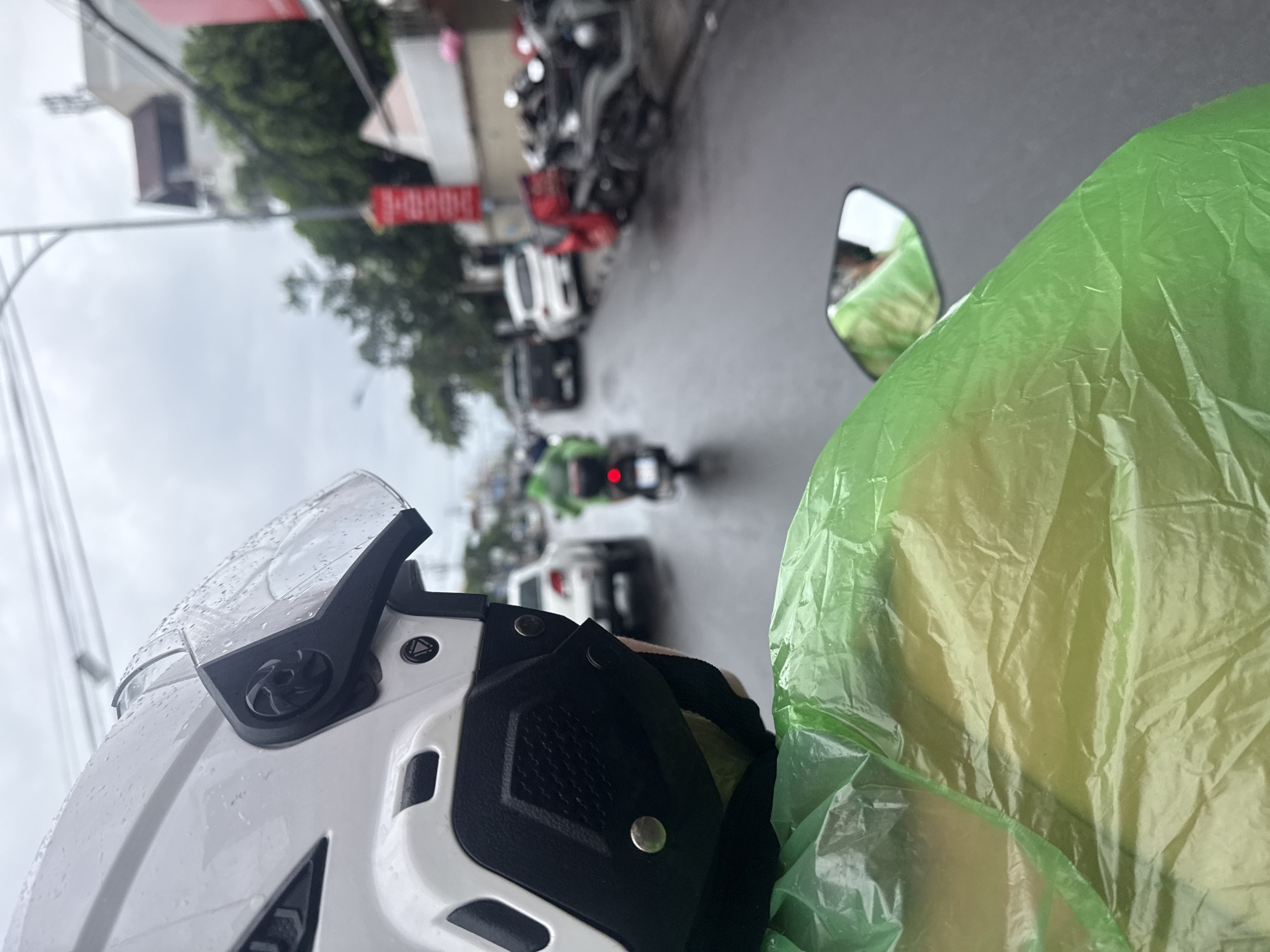The image captures a street scene in Nha Trang, Vietnam, during a downpour. The perspective is from the back of a motorcycle, with the rider wearing a white helmet and a bright green disposable raincoat. The road is wet and reflects the overcast sky, indicating rainy weather. Several vehicles are visible on the street, including cars, scooters, and a scooter with a green delivery bag, suggesting it might be a food delivery rider. Buildings and trees line the street, with red banners visible on one building, possibly indicating commercial establishments or events. The overall atmosphere is that of a typical rainy day in a Vietnamese city, with traffic moving along a wet road.