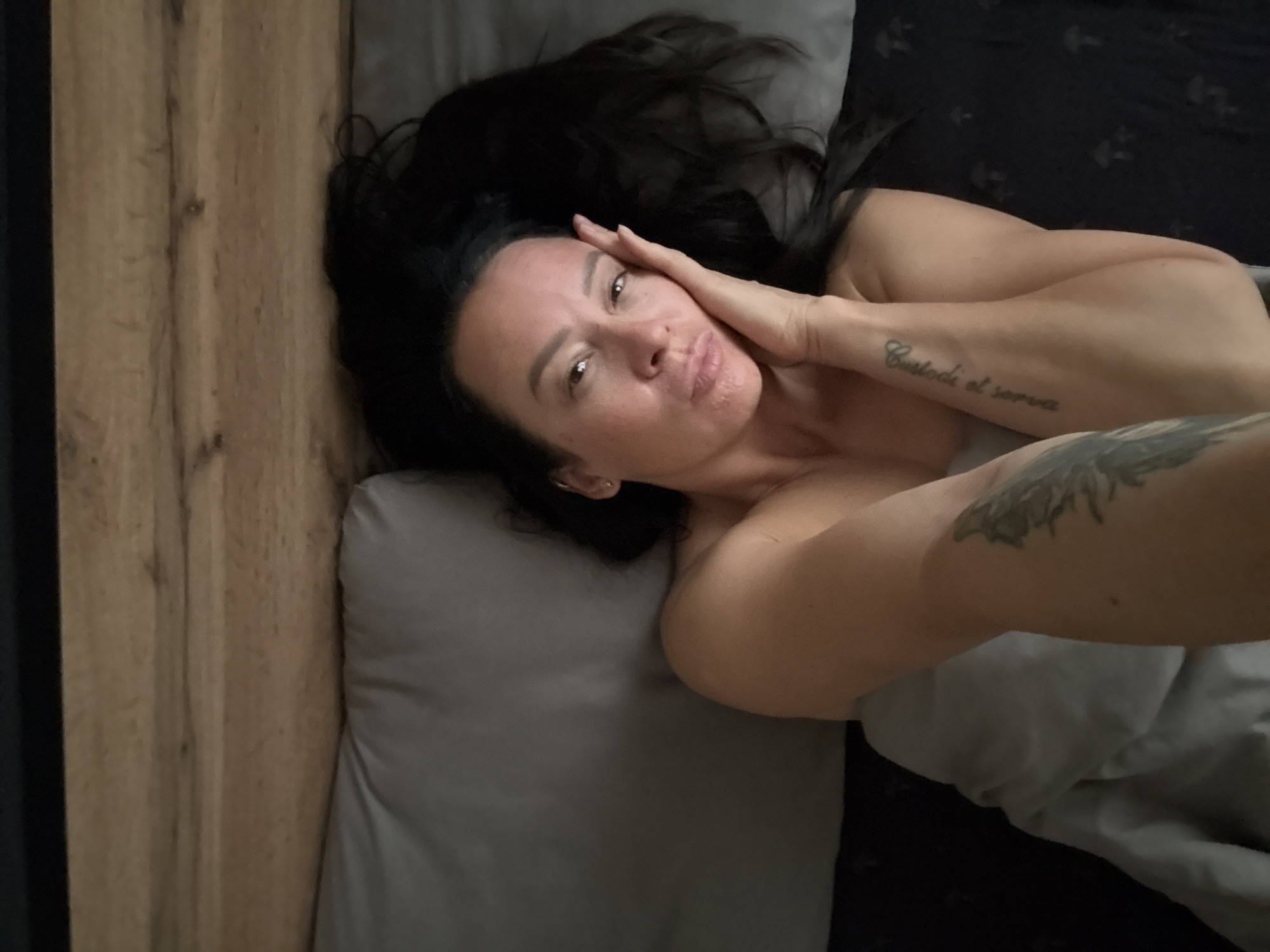 A woman with long dark hair lies in a bed, facing the camera. She is resting her head on her hand, with her eyes looking towards the viewer. She has a light-colored tattoo on her forearm that reads "Custodi et serva". The scene appears to be indoors, with a wooden headboard to the left of the frame and gray bedding. The lighting suggests it may be nighttime or an overcast day, as there are no strong shadows. The woman's expression is relaxed, and she seems to be posing for the photo. The image is cropped to focus on the woman and her immediate surroundings.