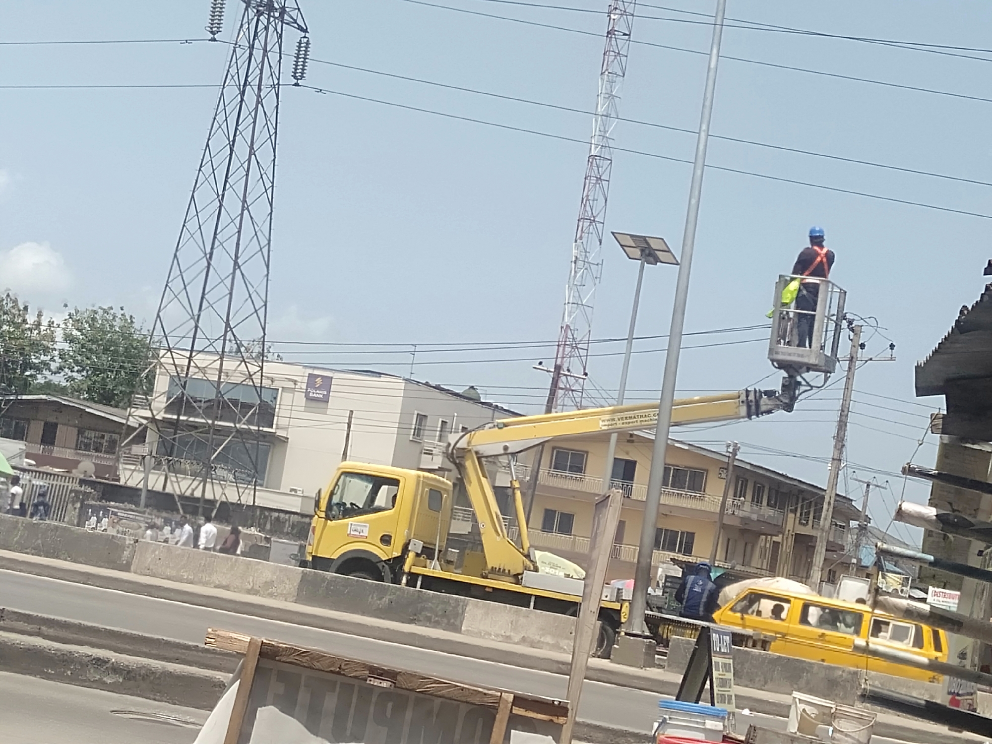 The image depicts a street scene in Shangisha, Nigeria, featuring a yellow crane truck with a worker in a safety harness and blue hard hat elevated in the crane's bucket. The worker appears to be performing maintenance on overhead power lines. Two lattice towers—a tall, dark metal structure and a thinner, white-and-red one—are visible in the background, along with a solar-powered street light. Several buildings are visible, including a bank bearing the Polaris Bank logo. A yellow mini bus with passengers is traveling down the road. A group of people is standing near the road. The road is divided by a concrete median. The scene is shot during daylight under a hazy sky.