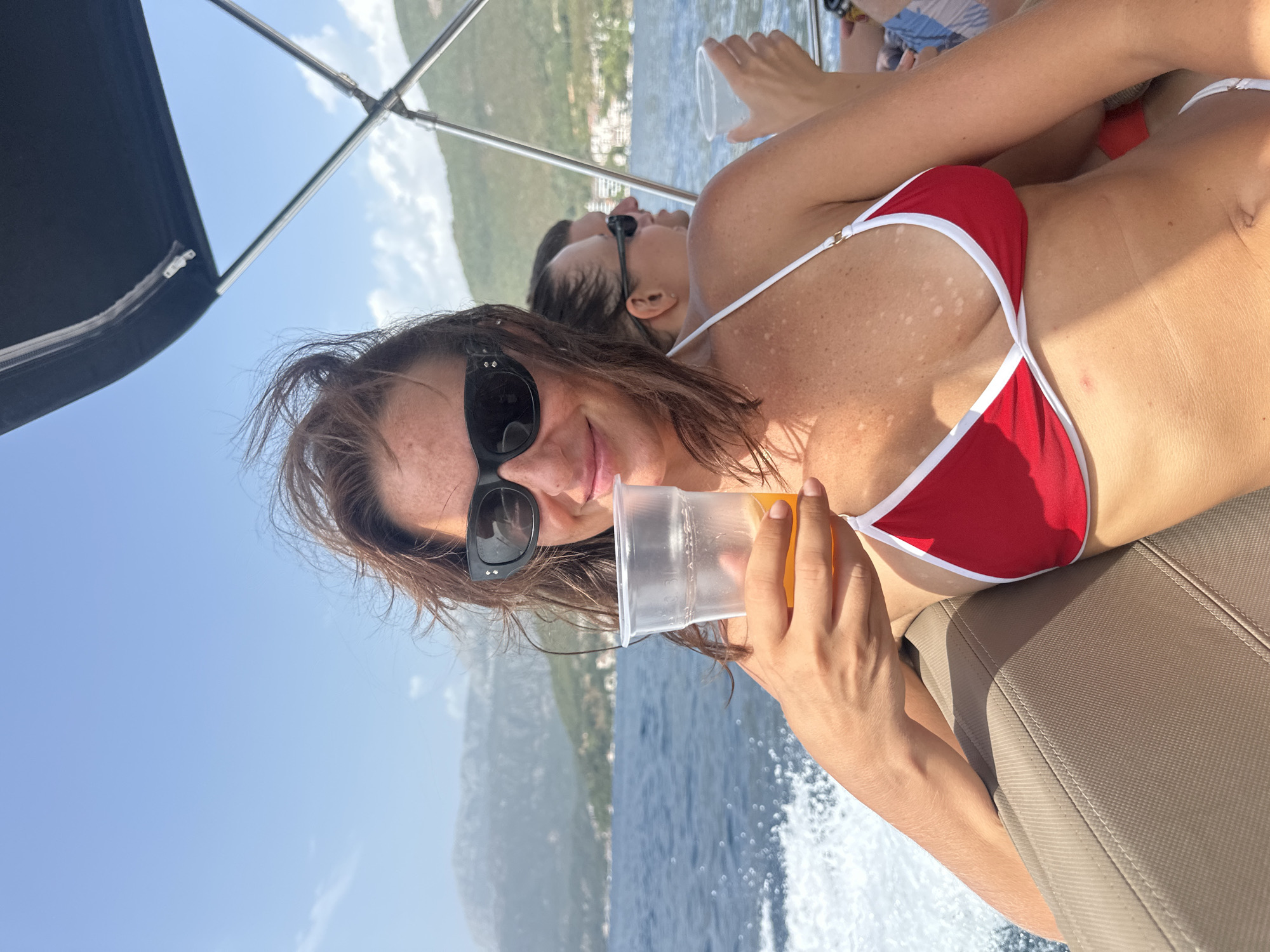 A woman with brown hair and sunglasses is holding a plastic cup of orange liquid while sitting on the deck of a boat. She is wearing a red bikini with white trim. Behind her, a man with sunglasses is also visible, and further back another person is seen holding a cup. The boat is moving through blue water, creating a wake. In the background, there are green hills with buildings, and a hazy mountainous landscape under a blue sky. The scene appears to be in Kumbor, Montenegro, given the distinct hilly coastline and the characteristic style of boats used in the area.