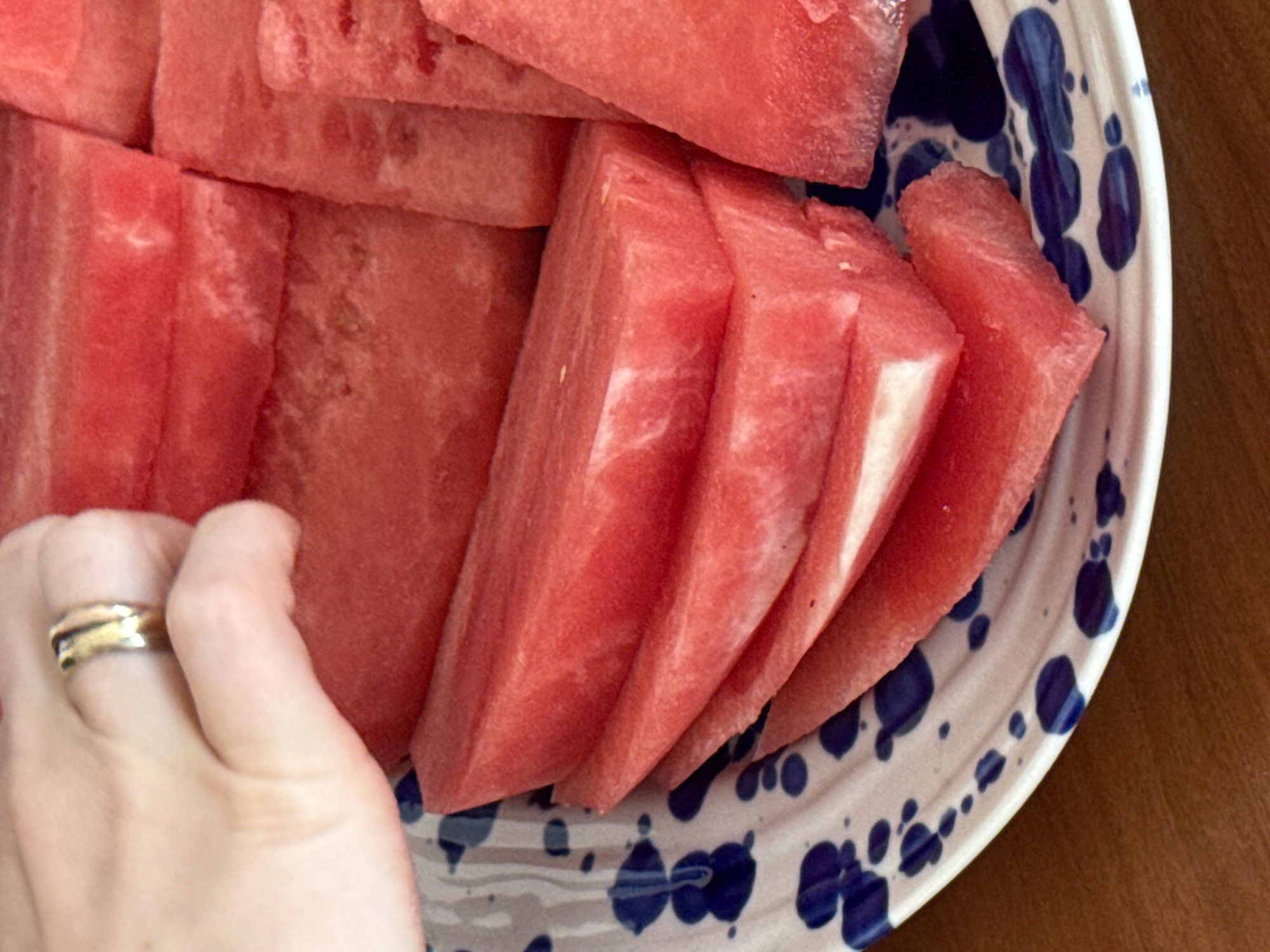 The image captures a close-up scene of a hand interacting with watermelon slices, situated in Tel-Aviv, Israel. A light-skinned hand, adorned with a gold-colored ring on the ring finger, extends from the upper left, positioned above several bright red watermelon slices. The watermelon, cut into various shapes including wedges and rectangles, displays a deep red flesh with subtle lighter veins and small, dark seeds. Some slices show the pale green-white edge of the rind. These slices are arranged on a white ceramic plate featuring an irregular, speckled blue pattern. The plate rests on a horizontal, warm brown, wood-grained surface, likely a table.