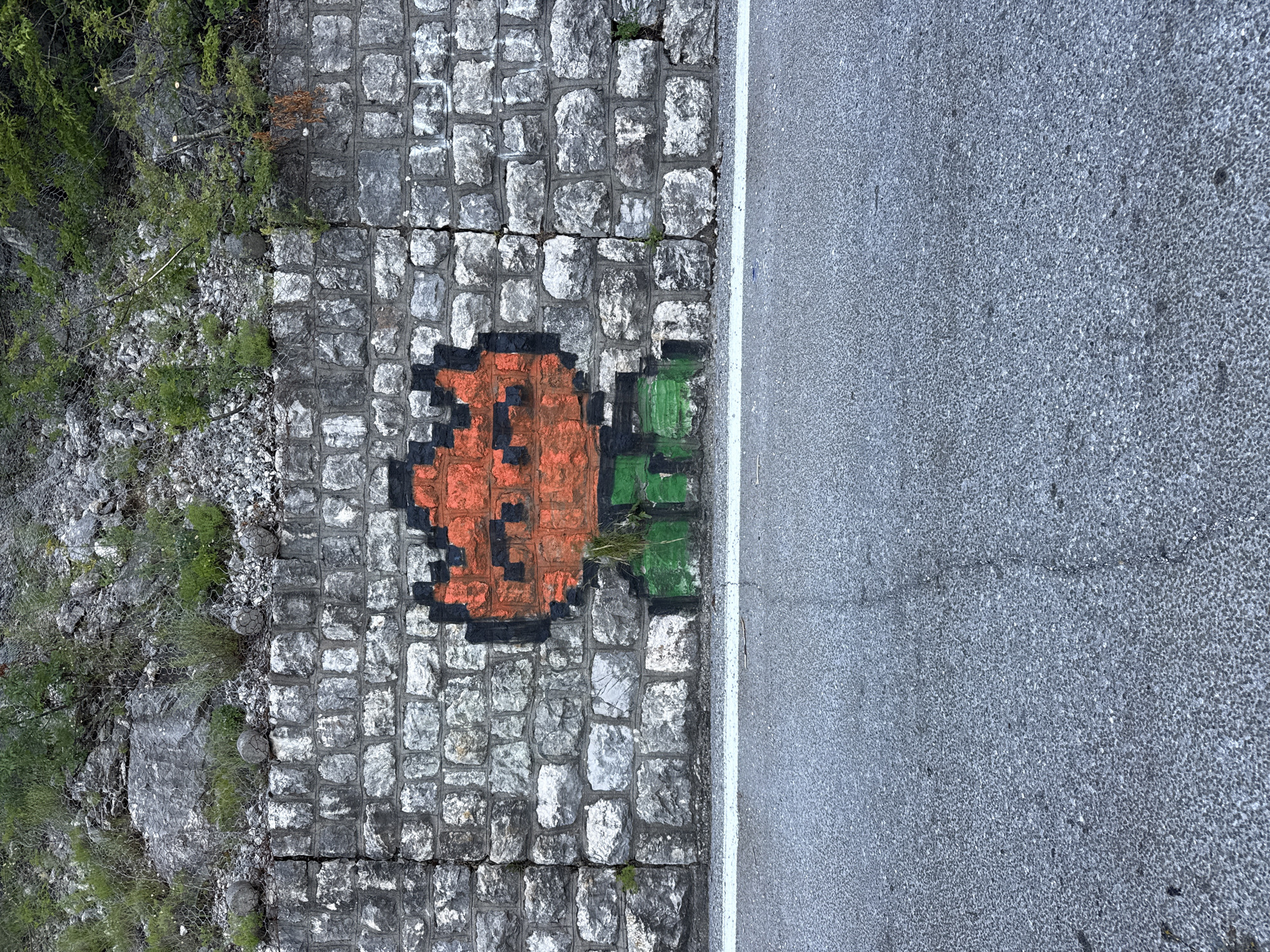 An orange and black pixelated alien-like figure is spray-painted on a grey stone wall. The figure has a square-shaped head with two black dots for eyes and a black outline. Below the head, it has green legs with black detailing. The wall is made of rough-hewn rectangular stones of various shades of grey and is cracked in several places. Above the wall, there is a rocky incline covered with sparse green vegetation and what appears to be a metal mesh fence. A white line marks the edge of a grey asphalt road in the foreground. The scene is located on the side of a road in Bukovik, Montenegro.