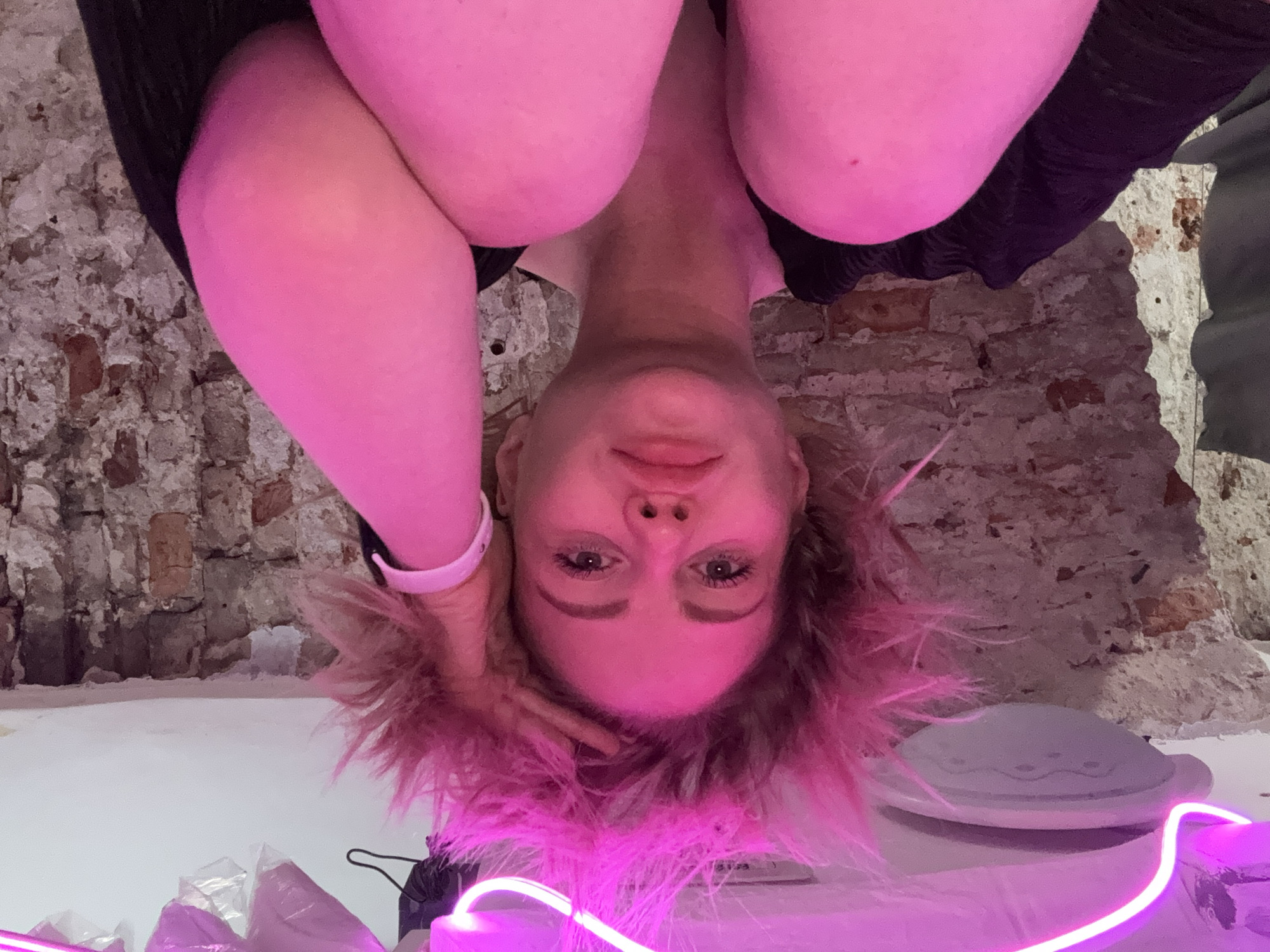 A person is upside down in front of a brick wall in what appears to be an interior space. The person has fair skin, light-colored hair that appears reddish or pink due to the lighting, and is wearing a dark-colored top. Their arms are positioned near their head. Below the person is a white surface with some objects, including what seems to be a small plastic bag and a section of a neon pink light. The brick wall behind them suggests the location might be an older building.
While the brick wall is suggestive of Saint Petersburg architecture, the scene's specific location within the city is undeterminable without more information.