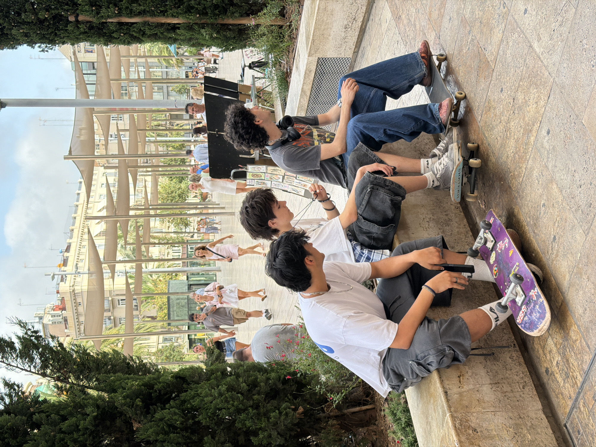 Three young males are sitting on a low, concrete ledge outdoors, with skateboards positioned on the ground in front of them. The individual on the left is wearing a white t-shirt and grey cargo shorts, with a purple skateboard featuring stickers placed in front of him. The middle individual has on a white t-shirt and dark grey shorts, with a blue and white skateboard at his feet. The person on the right is dressed in a grey graphic t-shirt and blue jeans, with a skateboard under his feet. They are all facing to their right and appear to be conversing. In the background, there is a wide pedestrian area with people walking, trees, and buildings with covered awnings overhead. The lighting suggests it is daytime.