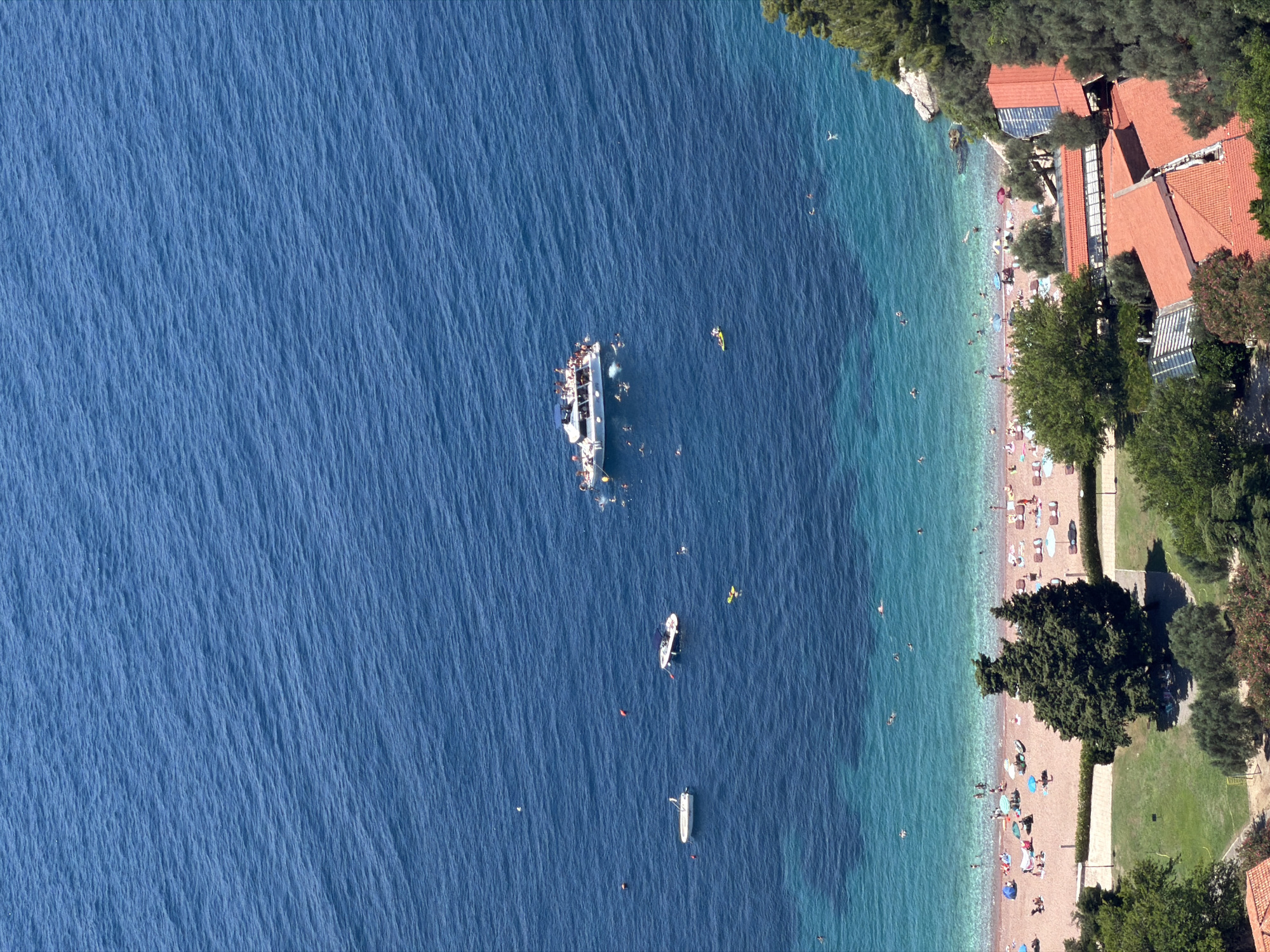 This is a high-angle, aerial photograph of a Mediterranean coastline in Sveti Stefan, Montenegro, during daylight. The deep blue sea occupies the upper portion of the image, with the turquoise shallows of the coast closer to the shore. A large white passenger boat, crowded with people, is anchored in the sea. Several smaller boats, including a white speedboat and a dinghy, are also present. Numerous individuals are visible swimming and enjoying the water.
The beach itself is sandy and populated with sunbathers resting on lounge chairs under umbrellas. A few trees dot the beach area. Behind the beach, a grassy area with more trees and several buildings with red tile roofs are visible. The overall scene depicts a popular tourist destination with active recreational use of the beach and sea.