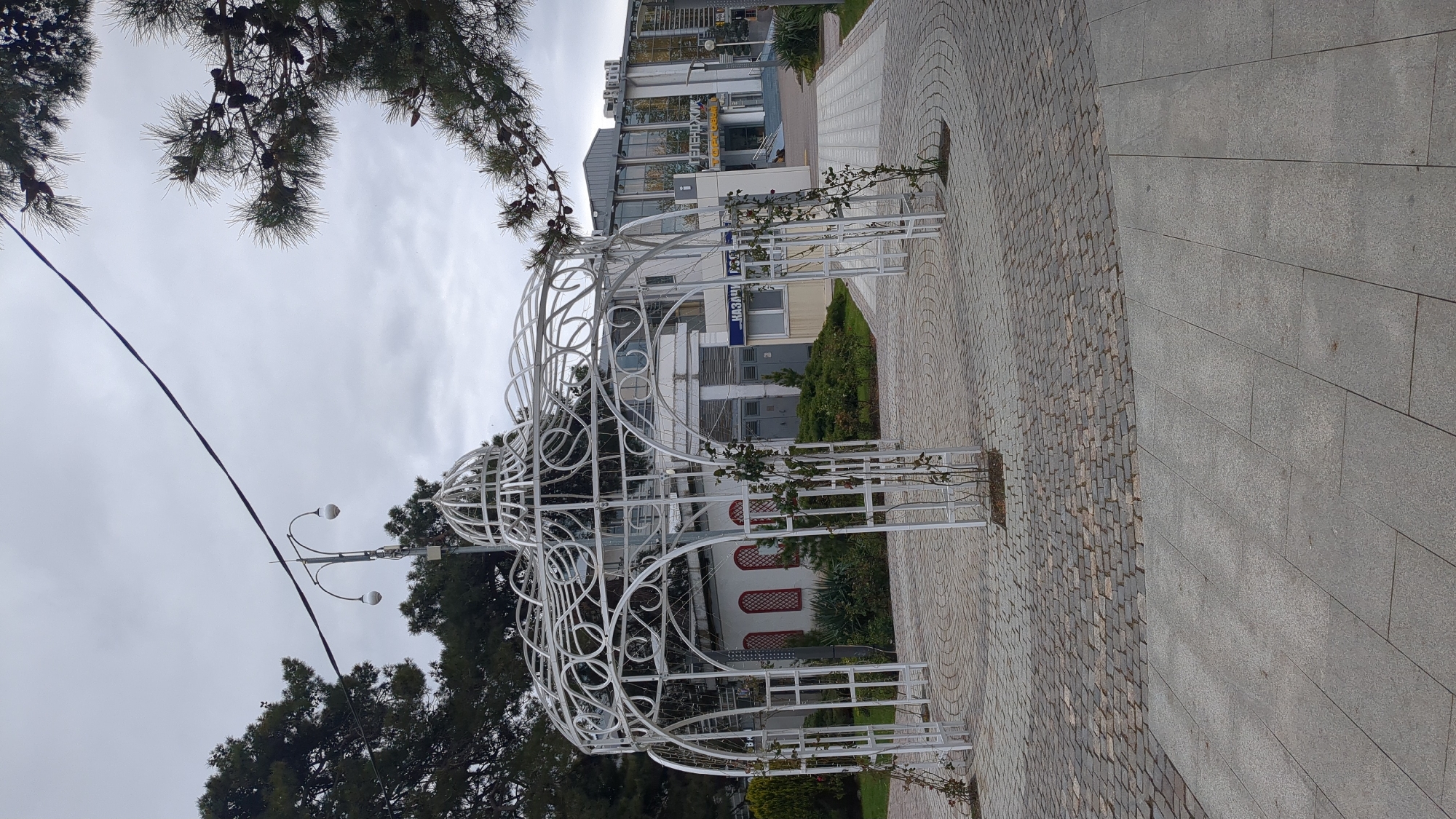 The image depicts a decorative white metal gazebo-like structure located on a paved walkway in Gelendzhik, Russia. The walkway is divided into sections of small cobblestones and large grey rectangular stones. The structure is partially covered with climbing plants, possibly roses. In the background, buildings are visible, including one with a sign in Russian that appears to say "МУЗЕИ". The sky is overcast. Trees are present to the left of the structure. A lamp post is visible behind the left side of the structure.