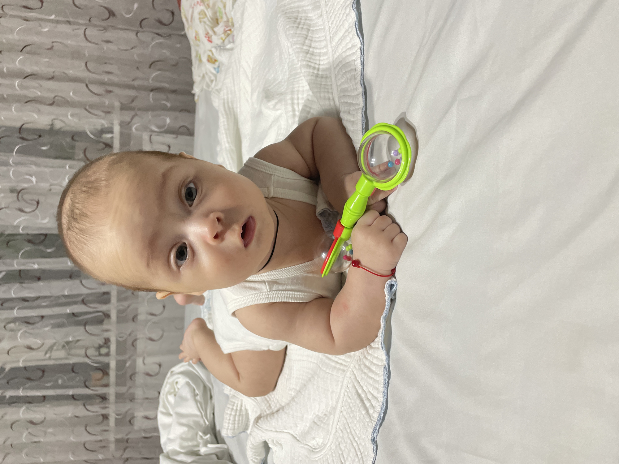 A baby is lying on its stomach on a bed, holding a green rattle. The baby is wearing a white tank top and has a red string bracelet on its left wrist. The bed has white sheets and a white blanket with a blue trim. In the background, there are sheer curtains. The scene appears to be indoors, likely in a bedroom. There is no information in the image that suggests the location is Fălești, Moldova.