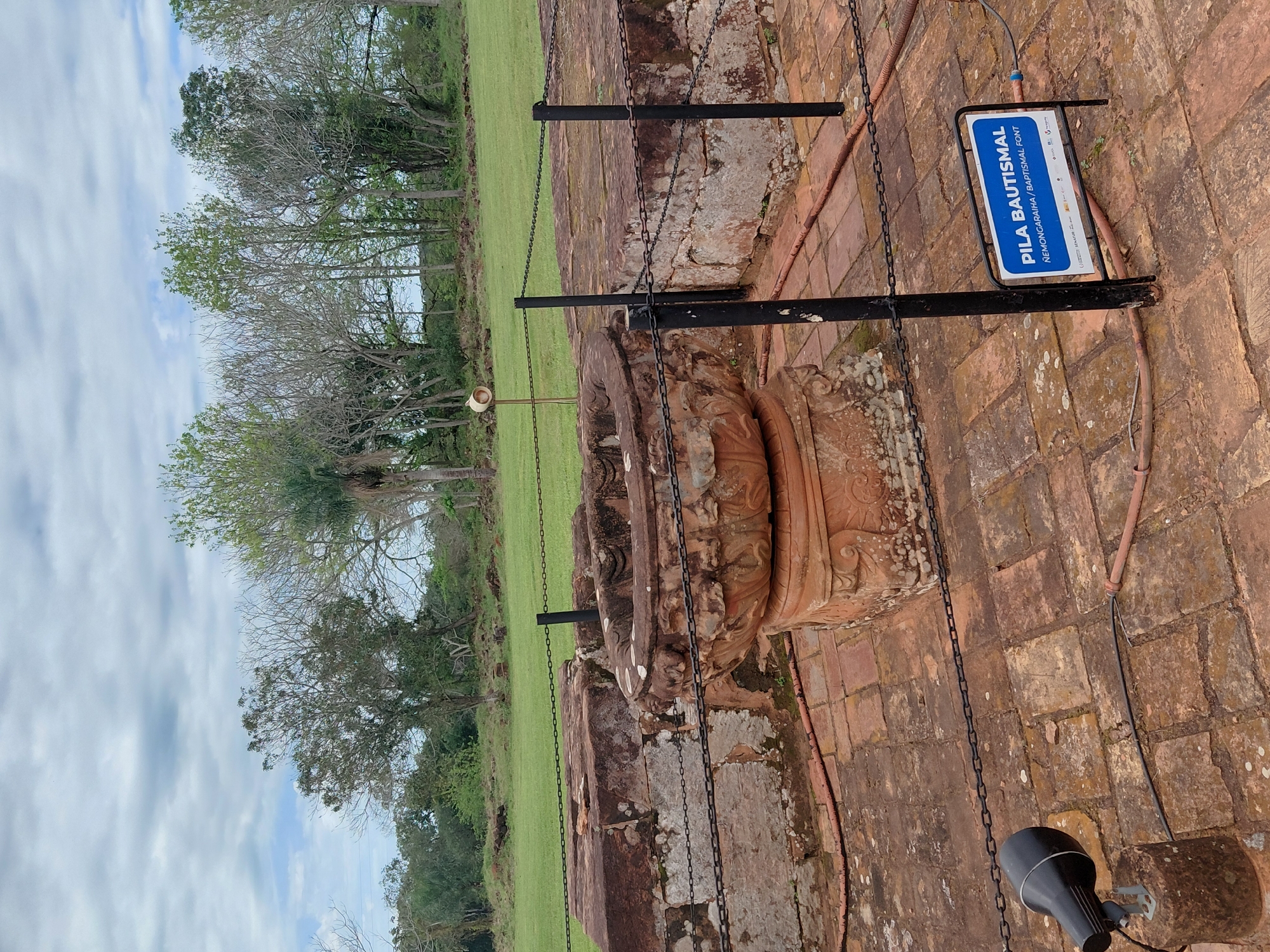 The image depicts an outdoor scene at the Jesuit Missions of La Santísima Trinidad de Paraná, located in Trinidad, Paraguay. In the foreground, a circular, ornate stone font, likely a baptismal font, sits on a pedestal of decorative terracotta. The font is surrounded by a brick-paved area, which is partially enclosed by a metal chain fence supported by black posts. To the left, a black spotlight is mounted on a concrete base, connected to a visible cable. A sign with blue and white lettering, reading "PILA BAUTISMAL / ÑEMONGARAIHA / BAPTISMAL FONT," is positioned near the fence on the right. In the background, large, weathered stone blocks, likely remnants of the historical mission, are visible, also partially sectioned off by the chain fence. Beyond the ruins, a lush green grassy field stretches towards a line of trees under a cloudy sky. The environment suggests a historical site preserved for visitors, with elements of preservation and information provided.