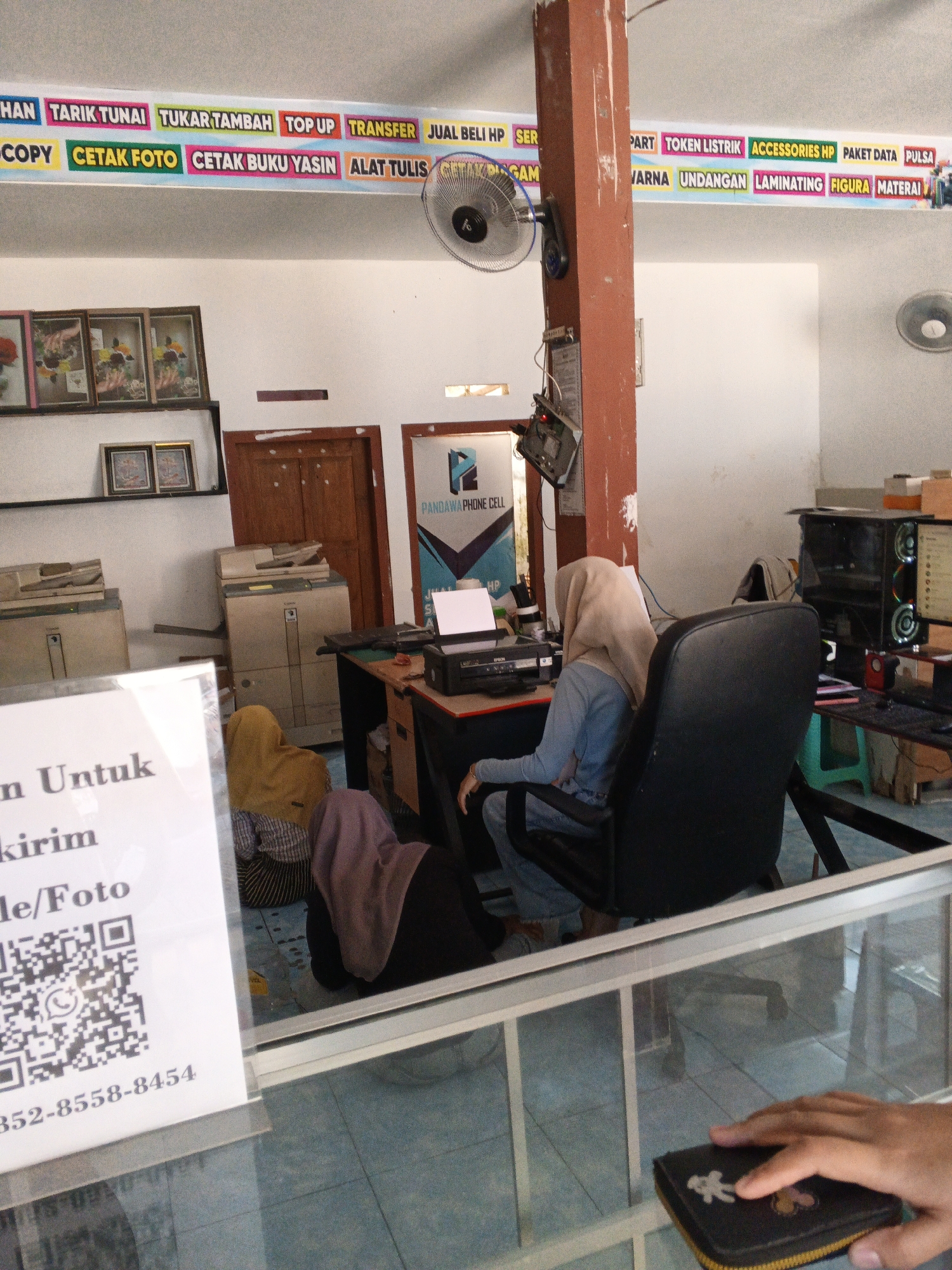 The image depicts the interior of a small business that appears to offer printing, copying, and potentially mobile phone-related services, likely located in Padang, Indonesia.
In the foreground, a hand is visible resting on a black wallet with a yellow zipper, placed on a glass surface. Behind this, a white sign with black text is partially visible, displaying "n Untuk" and "kirim" and "le/Foto". Below this text, a QR code is displayed, and further down, a phone number "352-8558-8454" is printed. This suggests the business might be involved in delivery or shipping services, or photo printing with contact information.
The middle ground of the image shows several people. A woman wearing a light brown hijab and a blue top is seated at a desk, looking towards a printer. To her left, another woman in a yellow hijab and a striped shirt is also seated, facing away from the camera. A third person, wearing a dark purple hijab and a dark garment, is seated on the floor or a low stool, also facing away. The desk itself has a printer, a computer monitor, and various papers and accessories.
Behind the seated individuals, several large office machines, likely photocopiers or printers, are visible. A wooden door is also present in the background, along with framed pictures on a shelf.
Above the scene, a banner with colorful lettering lists various services. These include "TARIK TUNAI", "TUKAR TAMBAH", "TOP UP", "TRANSFER", "JUAL BELI HP", "COPY", "CETAK FOTO", "CETAK BUKU YASIN", "ALAT TULIS", "PART", "TOKEN LISTRIK", "ACCESSORIES HP", "PAKET DATA", "PULSA", "VARNA", "UNDANGAN", "LAMINATING", "FIGURA", and "MATERAI". This comprehensive list confirms the business's diverse service offerings.
The environment appears to be an indoor retail or service space with tiled flooring and white walls. A fan is mounted on the wall to the right of the banner. The overall impression is a busy, functional service establishment.
