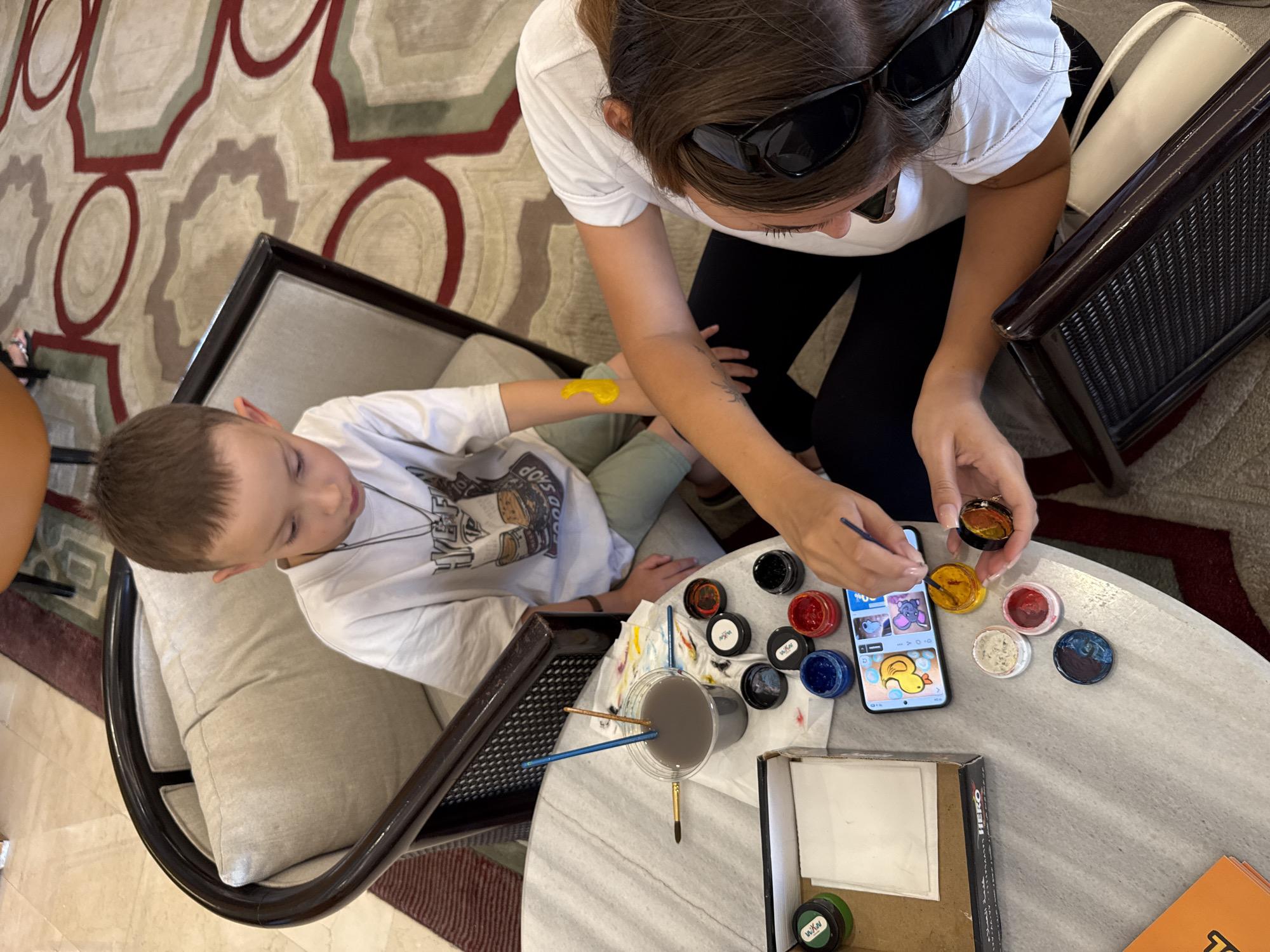 An elevated shot captures a mother and son engaged in face painting on a marble table in what appears to be a hotel lobby in Sharm El Sheikh, Egypt. The boy, seated in a dark-rimmed chair, wears a white t-shirt and green pants, with a partially painted yellow design on his upper left arm. His mother, seated across from him, wears a white top and black leggings, with sunglasses perched on her head. She is holding a paintbrush and appears to be painting on her son's arm. Scattered across the table are various containers of colorful face paint, brushes, a cup of grey liquid, a mobile phone displaying cartoon images, and a box labeled "HERO." The background reveals a patterned carpet with red, beige, and green designs, typical of hotel decor.