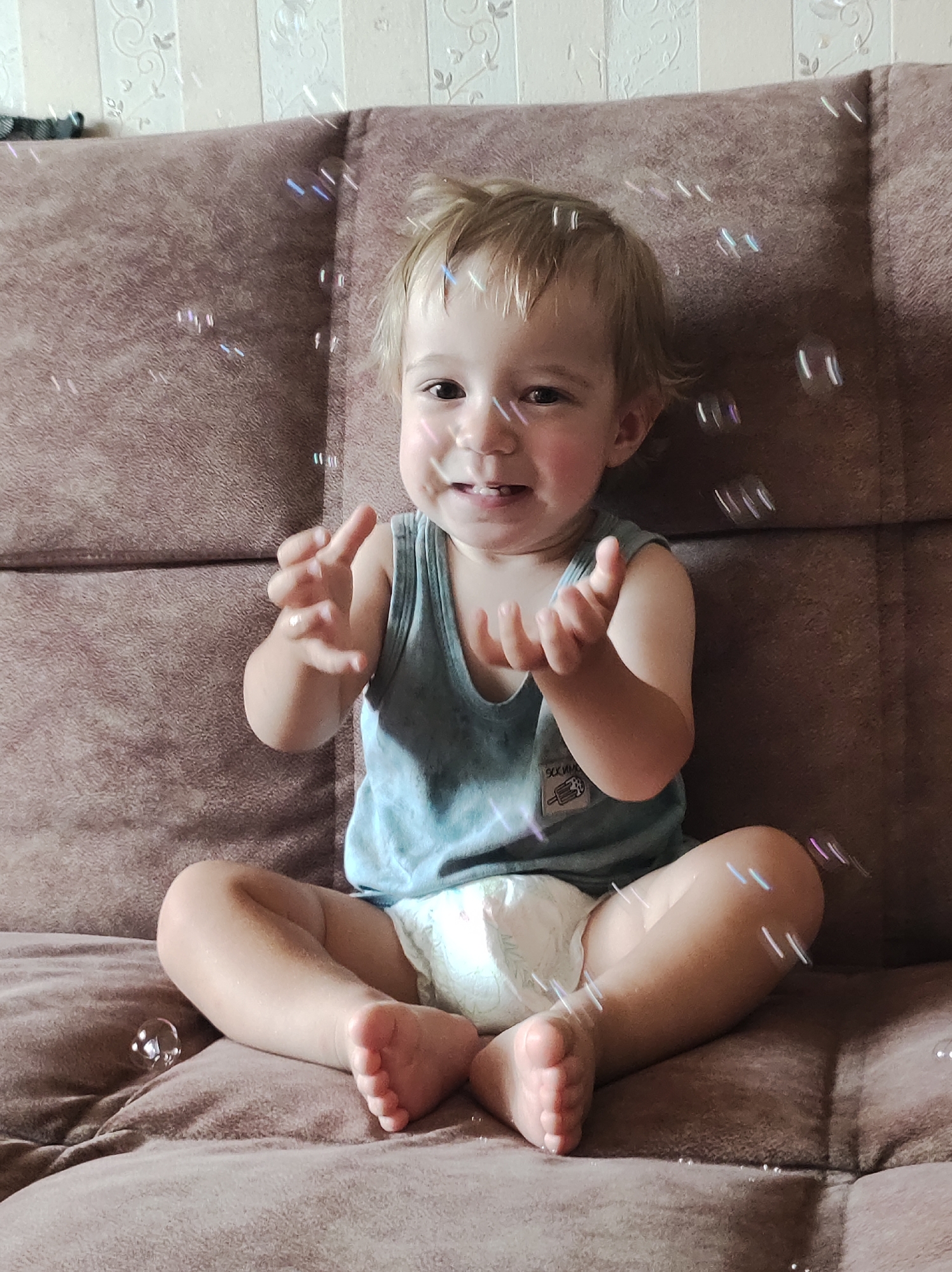 A young child sits on a brown sofa, playing with soap bubbles. The child, wearing a grey tank top and a diaper, is smiling and looking at the bubbles. The background consists of a patterned wallpaper. The image is taken indoors, likely in a residential setting. There is no visible indication of the city of Chaikovsky, Russia in the image.