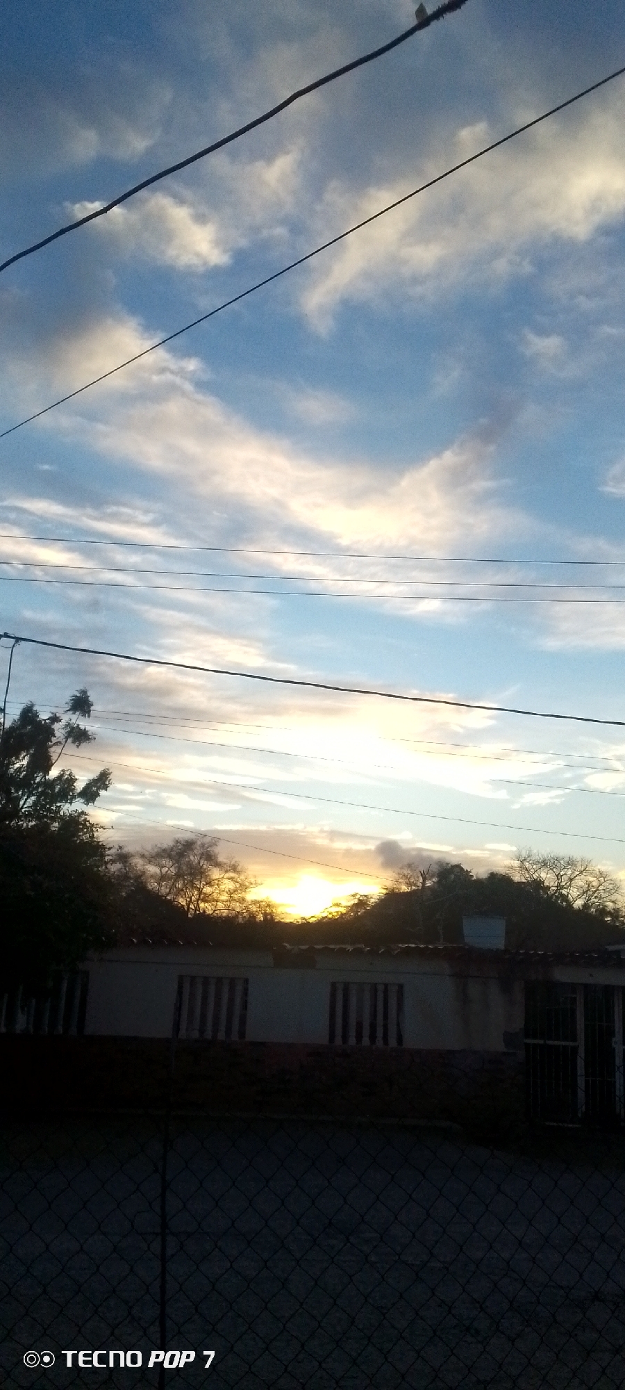 The image depicts a sunset in Altagracia, Venezuela. A long, one-story white building with vertical bars over the windows is partially visible in the mid-ground, obscured by trees. Above the building and trees, the sun is setting on the horizon. The sky is filled with scattered white clouds tinged with orange and yellow from the sunset. Power lines stretch across the upper half of the image. A chain-link fence fills the foreground. The bottom-left corner of the image includes the watermark "TECNO POP 7."