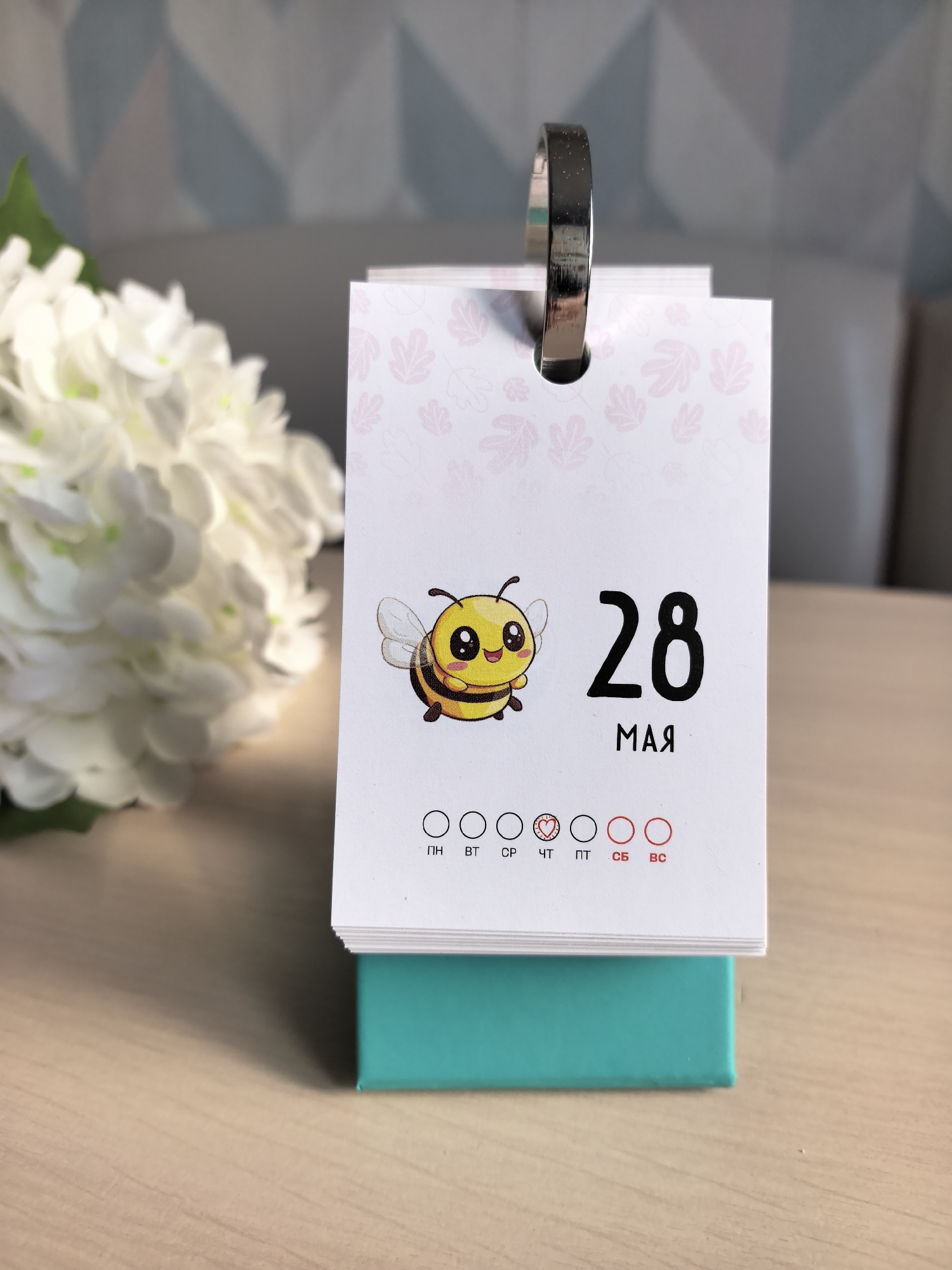 The image shows a tear-off calendar displaying "28 MAY" in Russian, adorned with a cartoon bee. The calendar is positioned on a teal base atop a light wood-grain surface. To the left is a cluster of white flowers. The background features a light gray sofa and a muted blue and white geometric pattern on the wall. Based on the calendar's language and the fact that "мая" is Russian for "May," the image is likely from Russia. However, the specific location of Фролы cannot be confirmed without further information directly provided in the image.