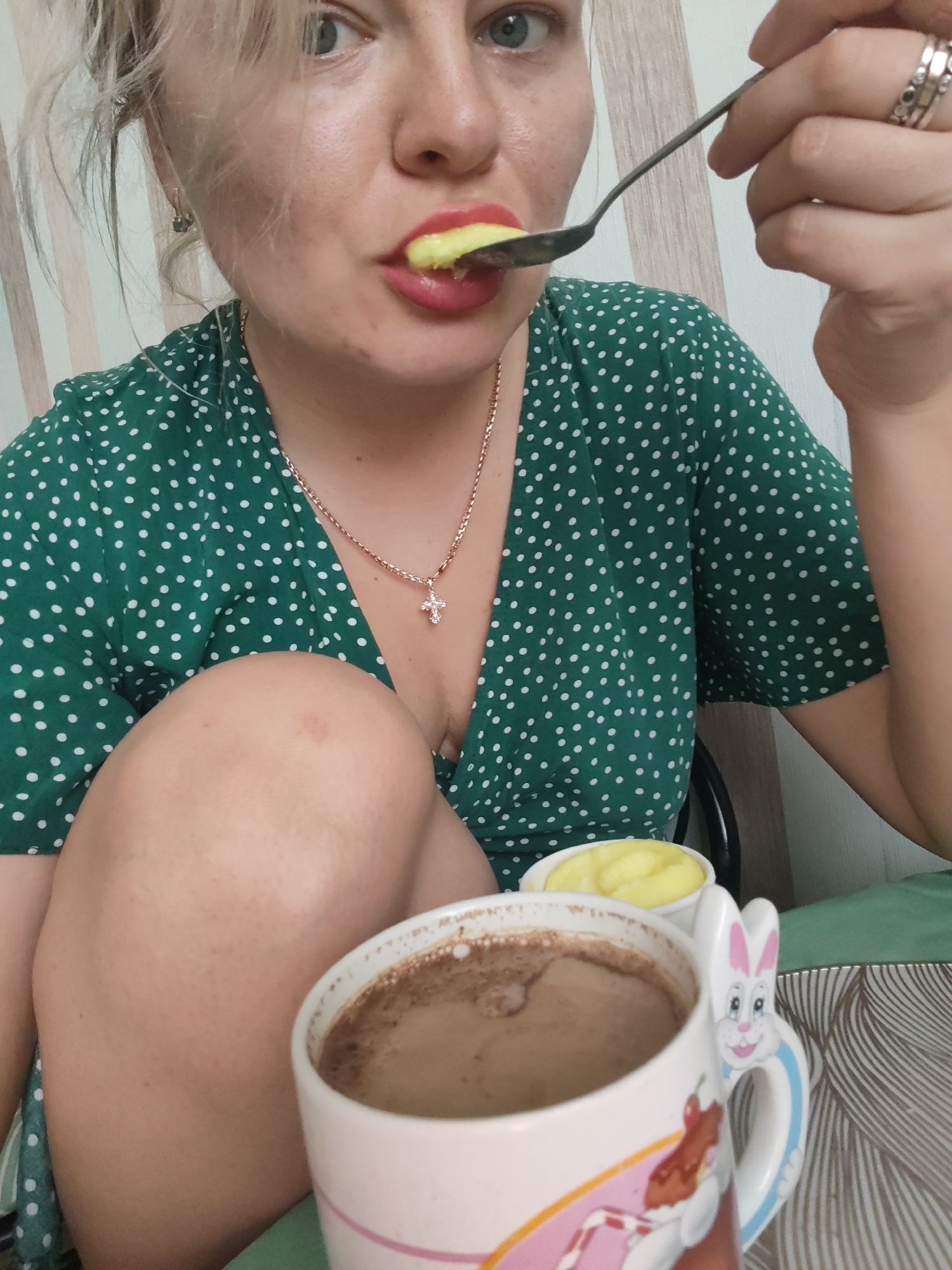 In an indoor setting, a woman with blonde hair, wearing a green polka dot dress and a cross necklace, is eating from a spoon. She has red lipstick on and is wearing rings on her fingers. In the foreground, there is a white mug with a bunny handle, filled with a brown beverage, likely coffee or hot chocolate. To the left of the mug, there is a small white bowl containing a yellow substance, possibly mashed potatoes or a dessert. The background shows a wall with vertical lines. There is no visible indication of the specific location within Izhevsk, Russia.