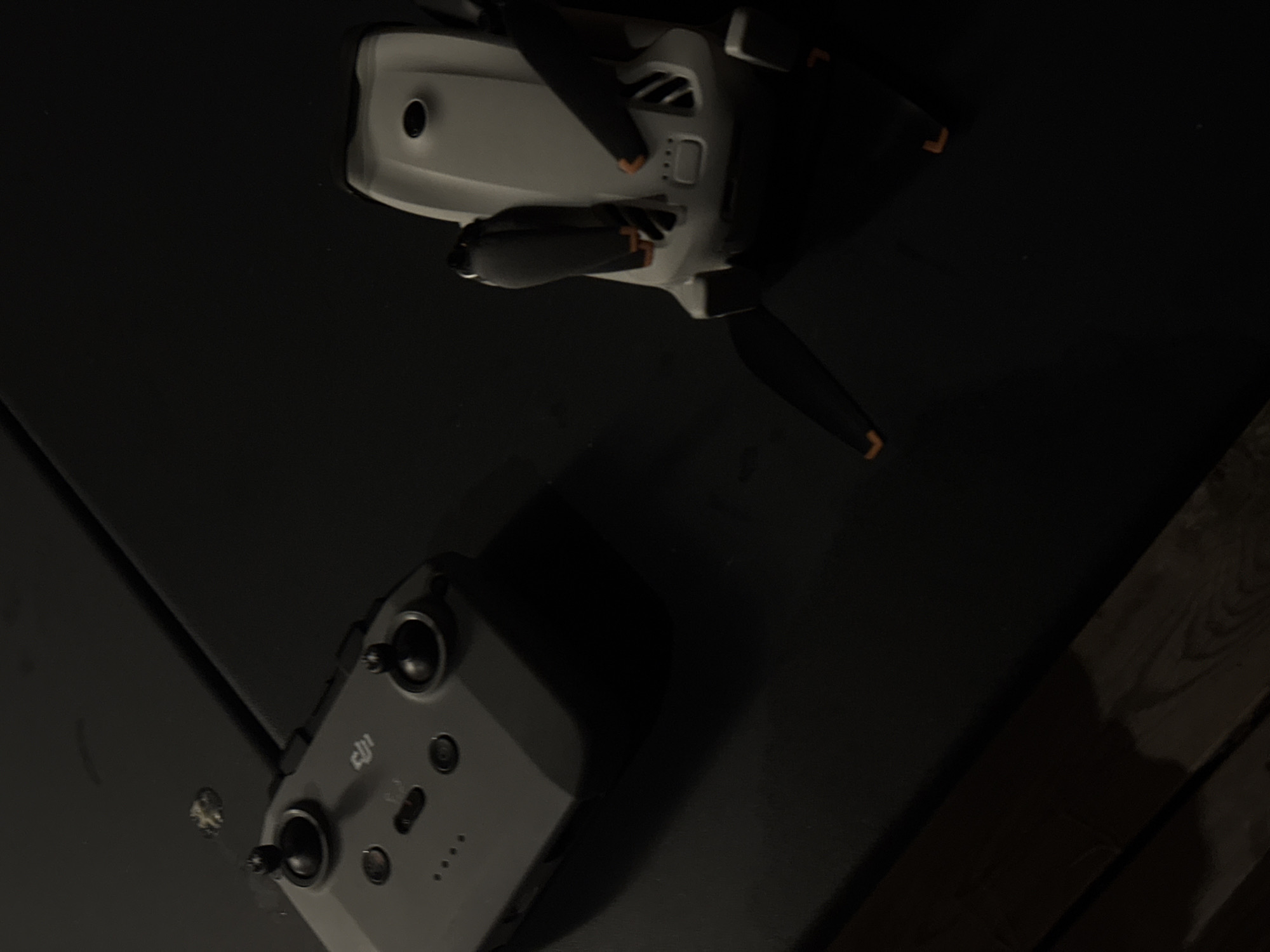 The image shows a DJI drone controller and a DJI drone lying on a dark, flat surface. The drone controller is grey with two joysticks and several buttons. The drone is white with black propellers that have orange tips. The scene is dimly lit, with shadows cast by the objects. The image does not contain any information about the location in the city of Vienne, France.