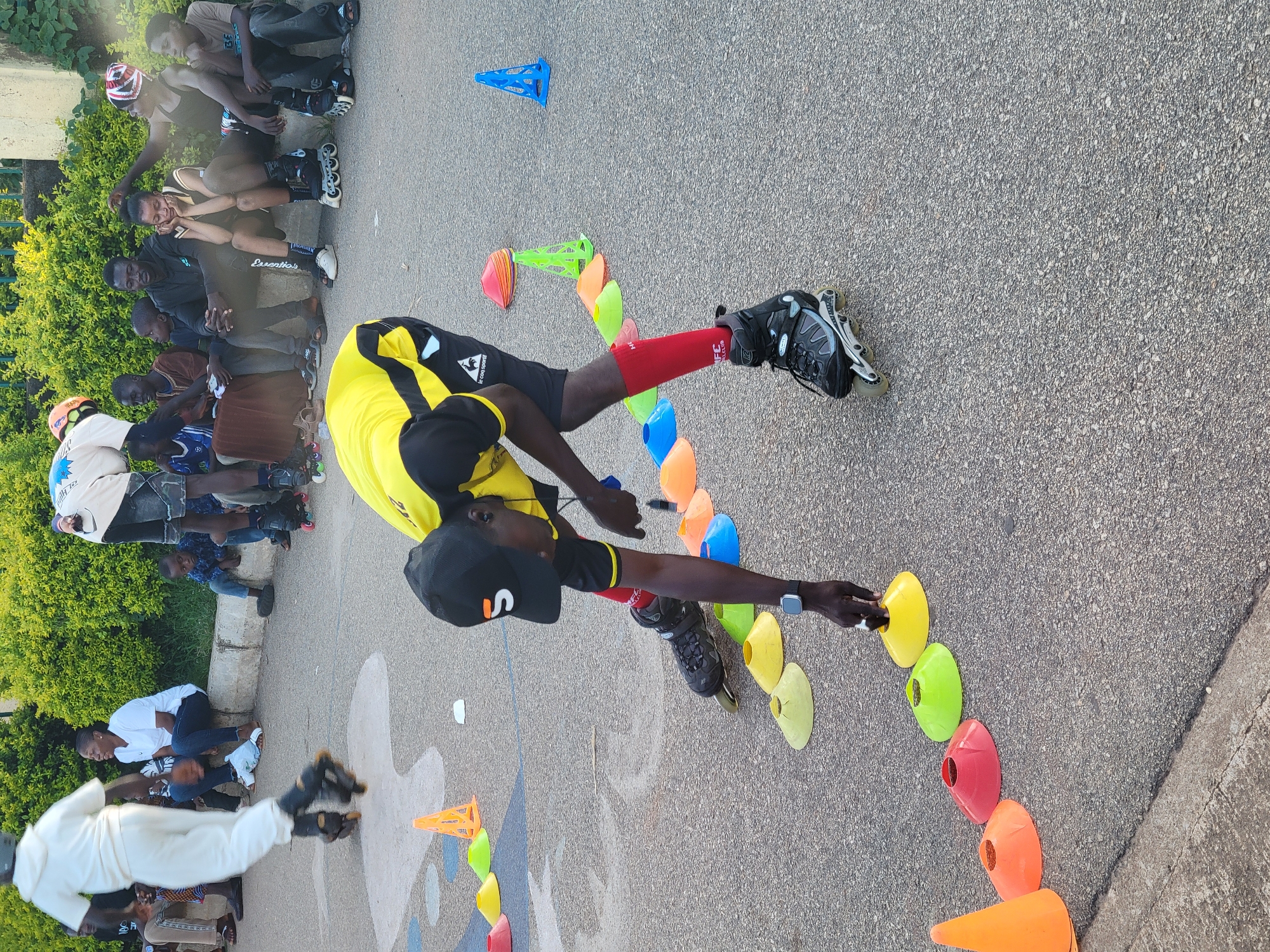 A group of people are gathered on an asphalt surface, possibly a paved area within a park or sports facility in Jos, Nigeria. In the foreground, a person wearing a yellow and black shirt, black shorts, red socks, and black rollerblades is bending down to touch a yellow cone placed on the ground. This person also wears a black baseball cap with a white "S" logo and a watch on their left wrist. Behind them, another person in white rollerblades and a white hooded garment is in motion, blurred due to movement.
Several colorful cones, including orange, yellow, lime green, and blue, are lined up on the ground in a formation, suggesting an obstacle course or training drill. In the background, a group of individuals are seated on a concrete ledge, some observing the activity. Among them are men, women, and children. One woman in a white t-shirt and jeans is seated, looking towards the rollerbladers. A young person in a blue t-shirt is also visible, as well as others wearing various clothing. Behind the seated group, a dense green hedge provides a backdrop. The overall scene depicts an outdoor activity involving rollerblading and cone drills, with spectators observing.
