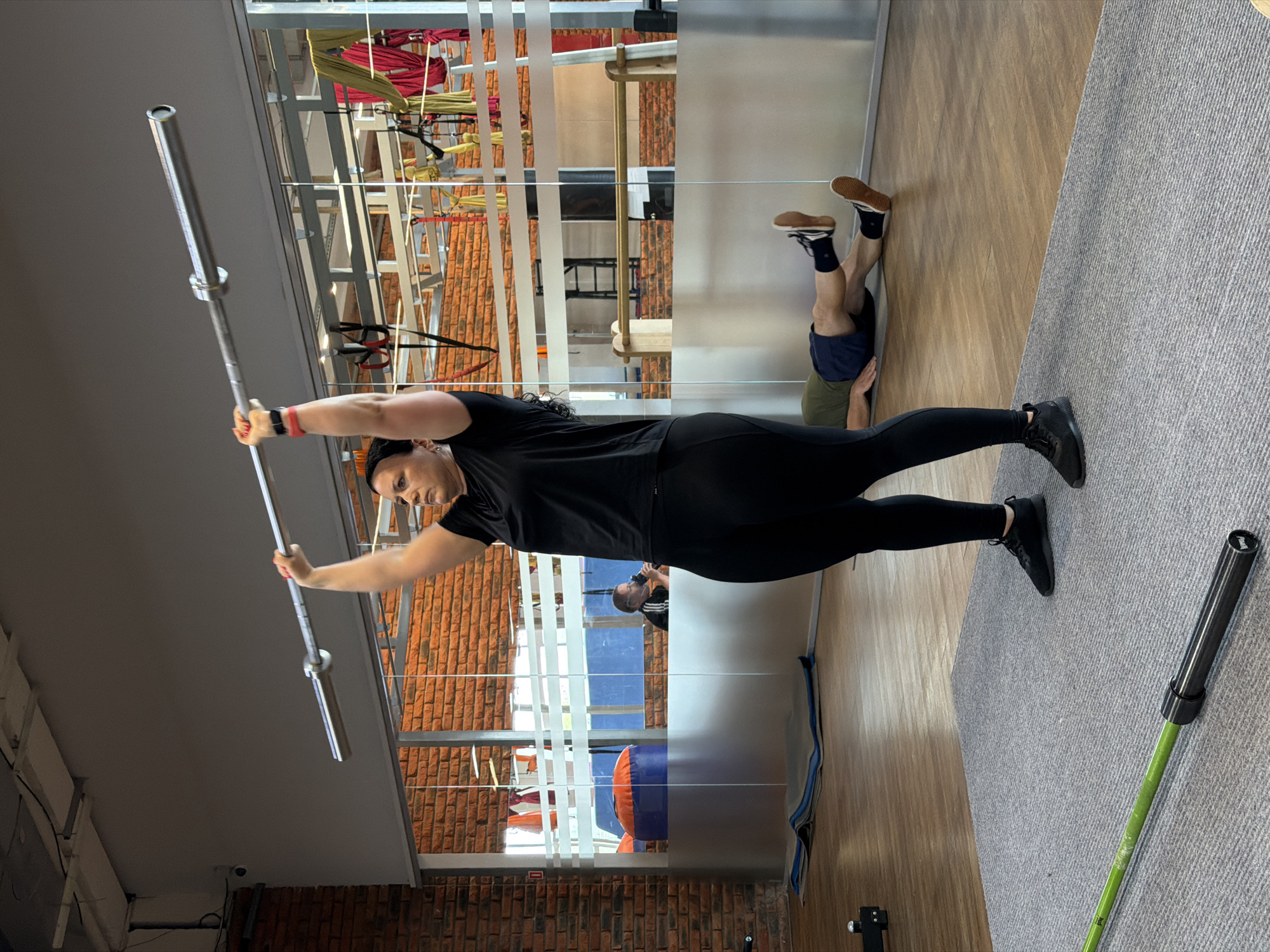 In a gym setting in Chaikovsky, Russia, a woman with dark hair is performing an overhead press with a barbell. She is wearing a black t-shirt, black leggings, and black athletic shoes. Her arms are extended upwards, holding the barbell steady. In the background, a man is lying on the floor with his legs bent and elevated, appearing to be engaged in an exercise. Another person can be seen in the reflection of a mirrored wall, seated and holding a water bottle. The gym environment features brick walls, wooden floors, and various gym equipment, including yoga mats and aerial silks hanging from the ceiling. The overall scene depicts a workout session in progress.