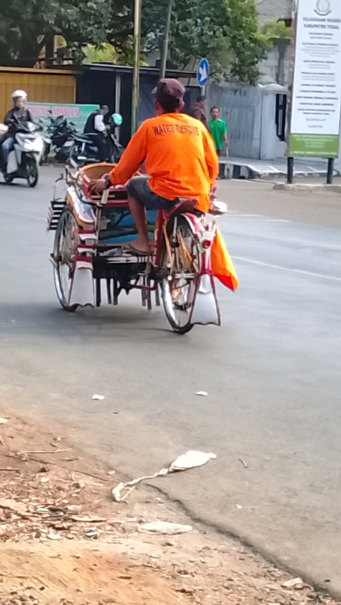 In Slawi, Indonesia, a man wearing an orange long-sleeved shirt with the text "WATER RESCUE" printed on the back is riding a pedicab on a paved road. The pedicab has a red and white frame and appears to be decorated with various colorful patterns. The man is wearing a dark cap and denim shorts, and his feet are bare, wearing flip-flops. In the background, there are trees, a building with a sign that reads "KELURAHAN DUWET," and several other people and vehicles, including motorcycles and a green shirted individual standing near a sign. The road surface is gray asphalt, and there is debris on the side of the road, including white plastic bags and a length of white fabric. The lighting suggests it is daytime.