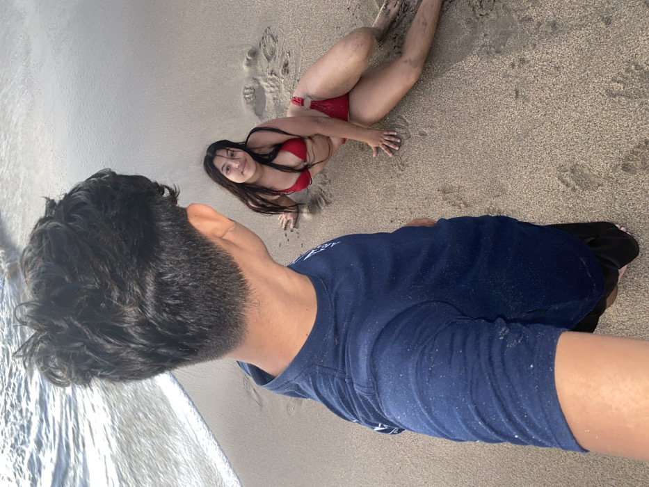 A man takes a selfie at the beach. He's wearing a navy blue long-sleeve shirt. Waves are visible in the background. A woman in a red bikini poses on the sand, looking up at the camera. Footprints dot the beach around her. The scene is sunlit and appears to be a casual beach outing.