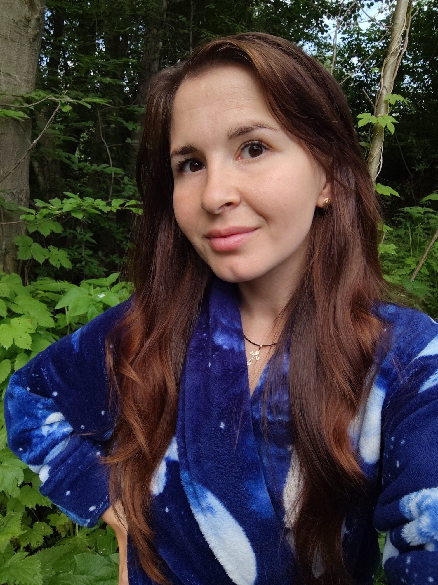A medium shot captures a young adult with fair skin and long, wavy brown hair smiling at the camera. She is wearing a plush, dark blue robe adorned with a white celestial pattern. A delicate gold necklace with a cross pendant is visible around her neck. Her left hand rests on her hip, while her right arm is bent. The background consists of lush green foliage and tree trunks, suggesting an outdoor, natural setting. The lighting appears to be natural daylight. The overall scene does not provide specific clues to pinpoint a location within the city of Фоки, Russia, as the environment is generic to many wooded areas.