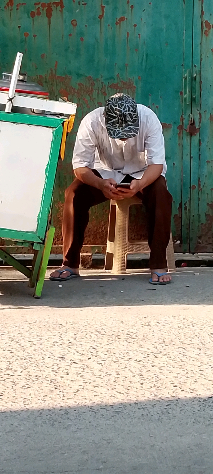 In Karangrahayu, Indonesia, a man wearing a white long-sleeved shirt, brown pants, and a patterned cap sits on a light brown plastic stool, engrossed in his smartphone. He is positioned in front of a weathered, rusty green metal door. To his left, a green-framed whiteboard is propped up on a stand. The ground is a textured, light-colored asphalt surface. The man's posture is relaxed, with his head bowed as he looks at the phone. His footwear consists of dark-colored flip-flops. The scene appears to be outdoors, with sunlight casting shadows on the ground.