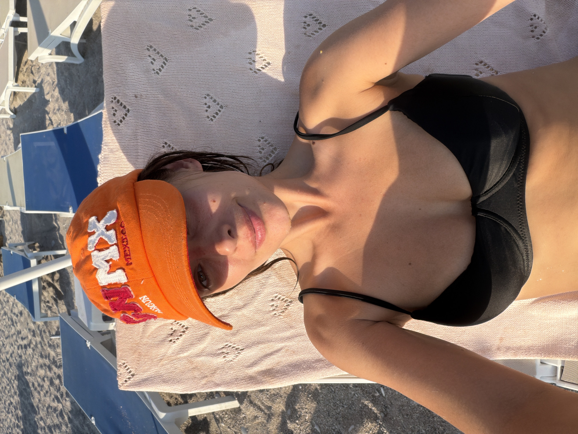 A woman lies on a beach chair on a sandy beach, wearing an orange baseball cap and a black bikini top. She is looking at the camera and smiling. The beach chair is covered with a pink towel. In the background, there are other beach chairs and umbrellas. The sun is setting, casting a warm glow on the scene. The image depicts a relaxing vacation scene at a beach in Fethiye, Turkey.
