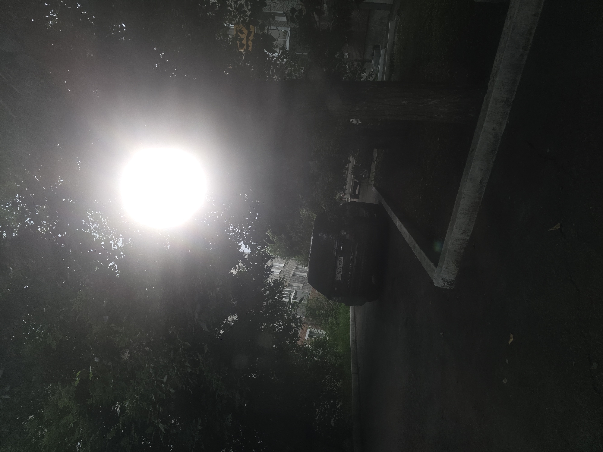 The image shows a bright, hazy sun shining through the leaves of trees, creating a lens flare effect. In the foreground, a dark-colored SUV is parked on a paved road next to a curb. The vehicle has a license plate with Russian characters and numbers. In the background, a multi-story apartment building with visible windows is present, surrounded by greenery. The overall scene suggests an outdoor urban environment, likely a residential area in Ulan-Ude, Russia, during daylight hours.