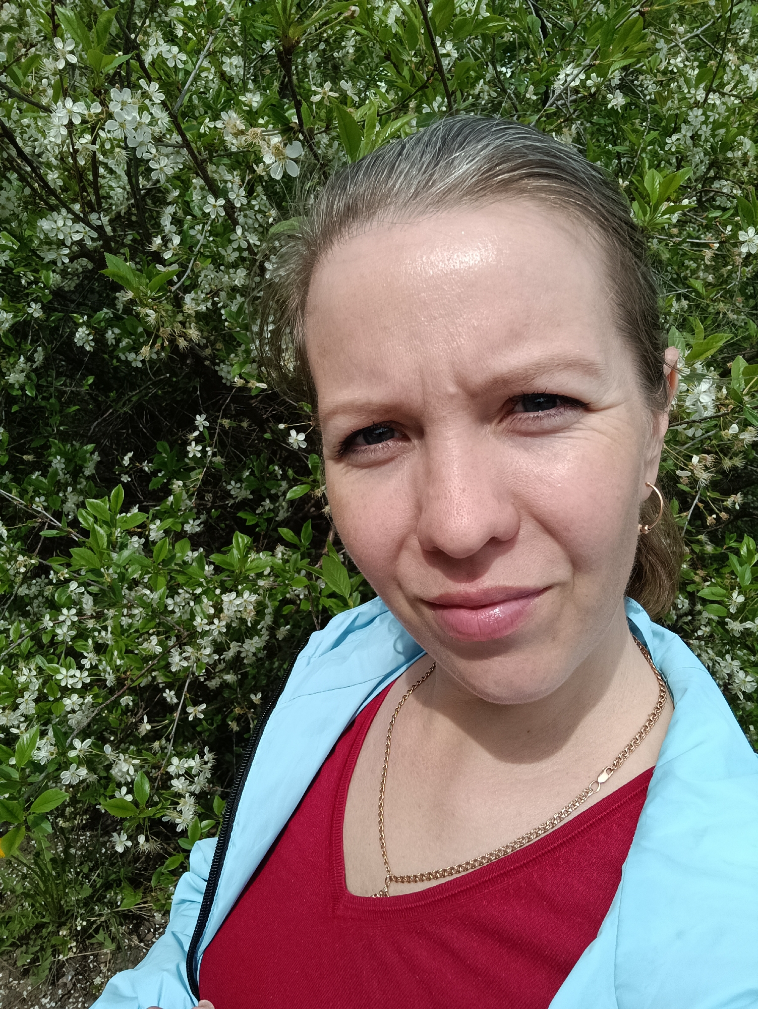 A medium close-up shot shows a middle-aged woman posing for a selfie or photo. She has light skin, graying blonde hair pulled back, and is wearing a red shirt and a light blue jacket. A gold necklace hangs around her neck. Behind her, there is a dense bush with small white flowers and green leaves. The location is likely a public park or garden setting in Chaikovsky, Russia.