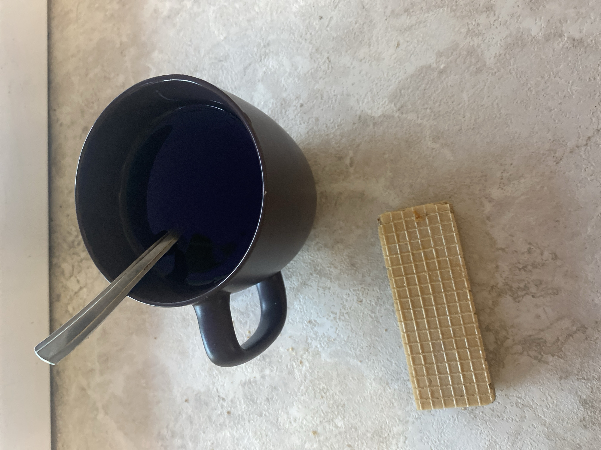An overhead view captures a still-life arrangement featuring a beverage and a snack on a light-colored, textured countertop. A dark-colored, possibly navy blue or black, ceramic mug is positioned on the left, containing a striking dark blue liquid. A silver spoon rests inside the mug, with its handle extending towards the bottom left of the frame. To the right of the mug lies a rectangular wafer cookie, characterized by its grid-like pattern and light beige color, suggesting a crispy texture. The background shows the mottled surface of the countertop, with a narrow, vertical white strip, likely part of a wall or cabinetry, visible along the far left edge. The scene suggests a quiet moment of refreshment, possibly a daytime snack or drink in an indoor setting like a kitchen or dining area in Chaikovsky, Russia. There are no visible people, specific location markers within the image itself, or discernible text.
