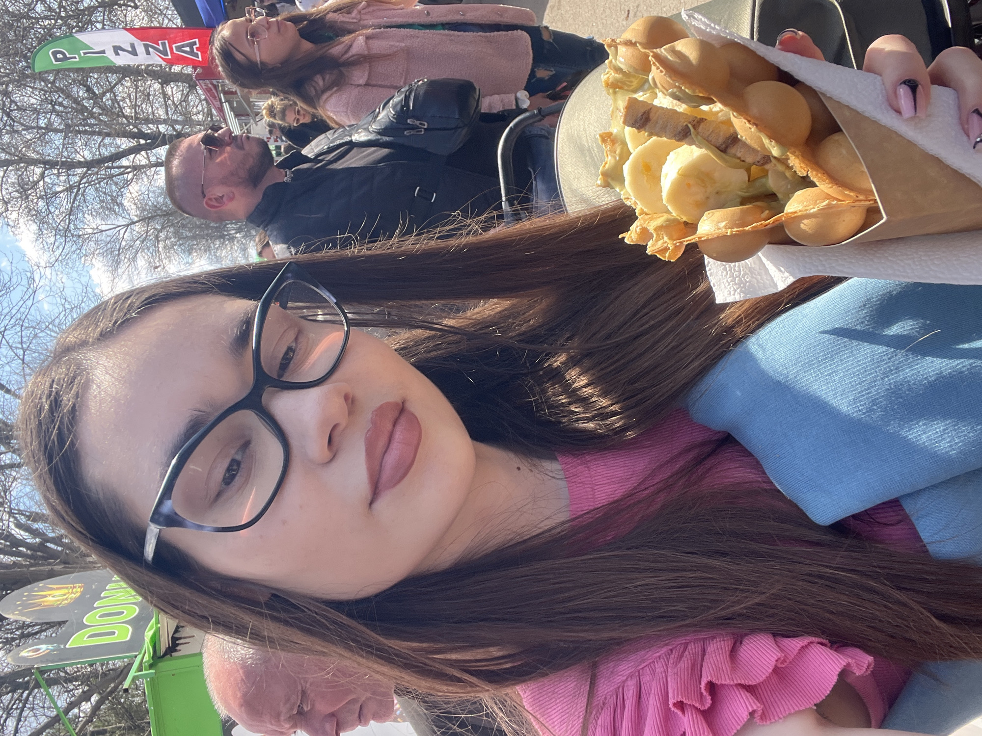 A young woman is holding a bubble waffle cone filled with banana slices and other fillings. She is wearing glasses, a pink top, and a blue sweater. In the background, there are several people, a Pizza flag, and a Donut sign. The exact location in Provadia, Bulgaria, cannot be determined from the image.