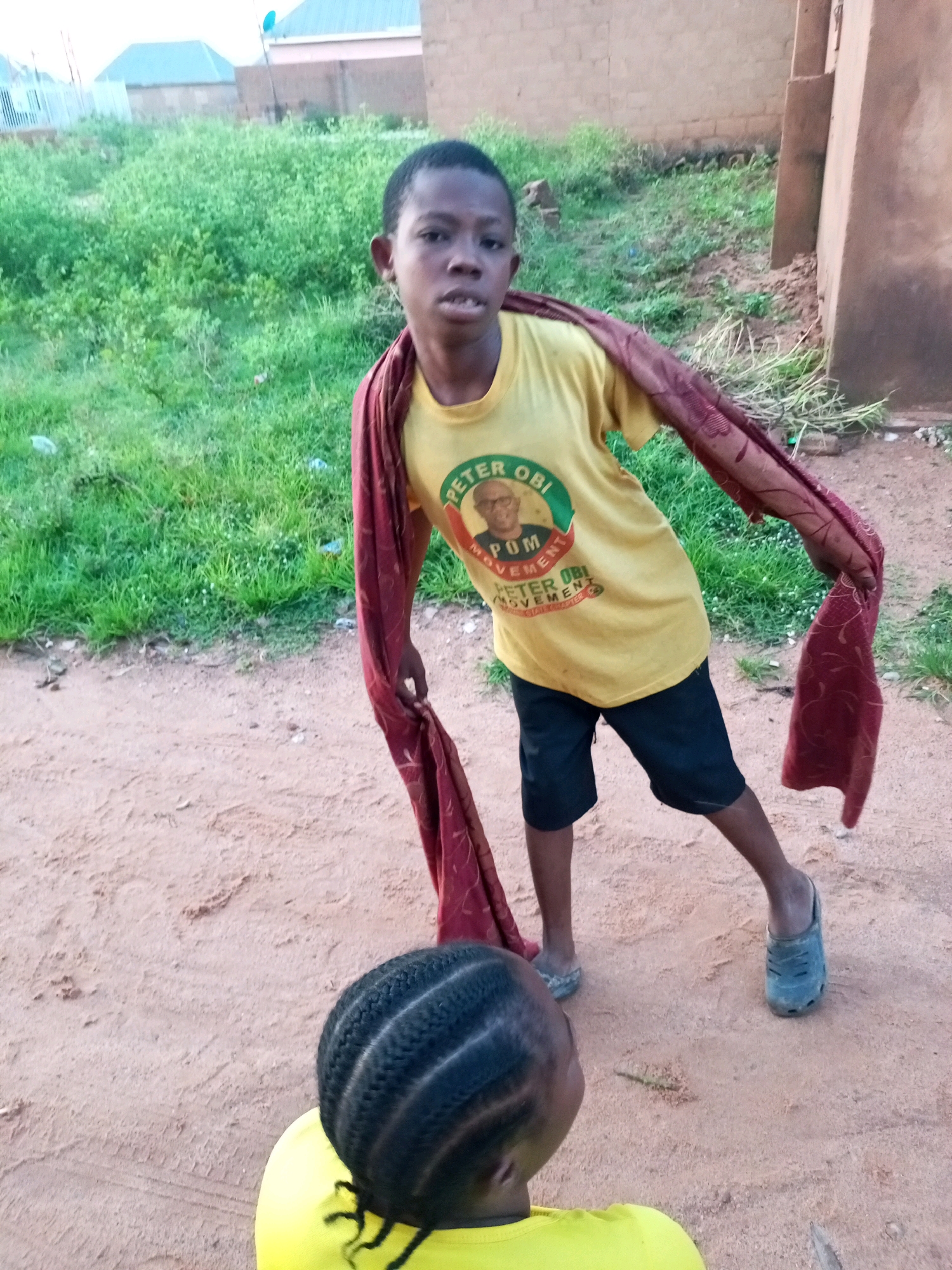 An elevated shot captures two young individuals in an outdoor environment. The primary subject, a male child, stands facing the camera, wearing a yellow t-shirt with the imprint "PETER OBI MOVEMENT" and a portrait of Peter Obi. He also wears black shorts and blue clogs. A maroon cloth with a floral pattern is draped over his shoulders and held in his right hand. He appears to be in motion, with his left leg bent and his right leg extended. To his left, partially visible in the foreground, is another child, from behind, wearing a yellow shirt, with braided dark hair. The background consists of dry, sandy ground, patches of green grass, and a brick building. The scene suggests an informal gathering or street activity. This location is identifiable as the city of Tempure, Nigeria, given the context provided.