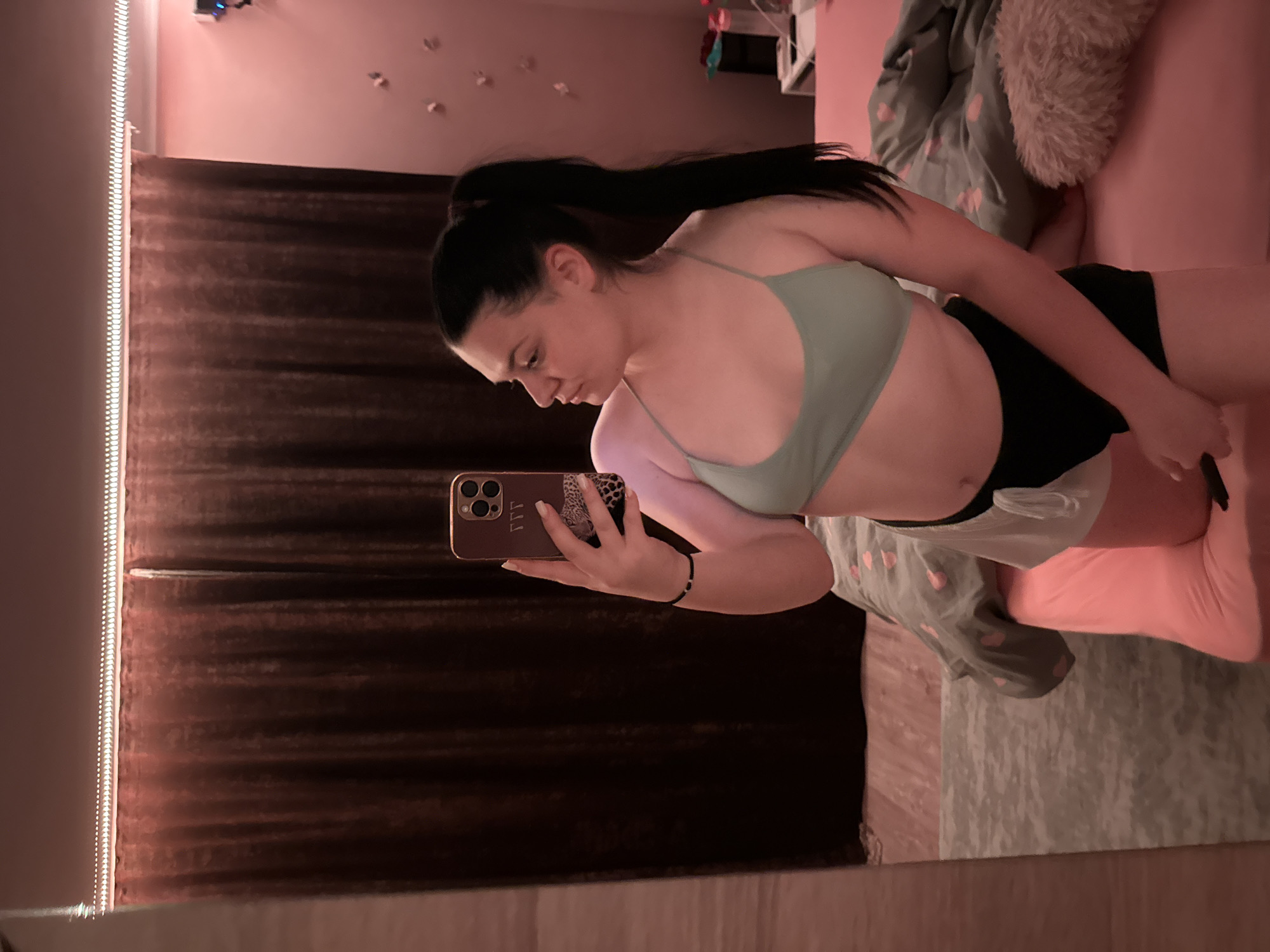A young woman with dark hair pulled back in a ponytail takes a mirror selfie in a bedroom. She is wearing a mint green sports bra and black shorts. The room has dark brown curtains and pink lighting. A bed with pink and grey bedding is visible behind her. The floor is wooden and a rug is on the left side of the image. There is a phone in her hand with a leopard print case. The woman is looking away from the camera, towards the right side of the room. The image does not provide any information to determine a specific location in the city of Rogosh, Bulgaria.