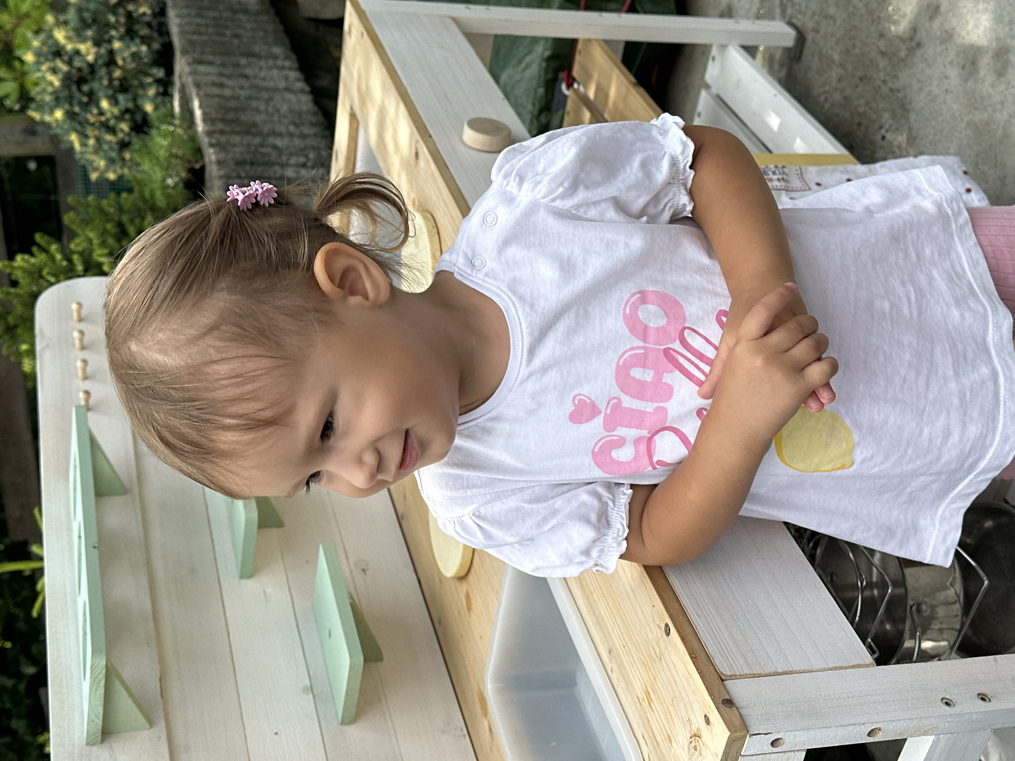 The image shows a young girl with light brown hair pulled back with pink flower clips, wearing a white t-shirt with pink lettering and a yellow circle. She is standing in front of a wooden play kitchen with light green shelves. The play kitchen has a gray sink and a wooden countertop. In the background, there are various green plants and a concrete planter. The ground appears to be concrete or stone paving. The girl's hands are clasped in front of her. The scene is outdoors. There is no specific information about the location in the city of Wisła, Poland within the image itself.