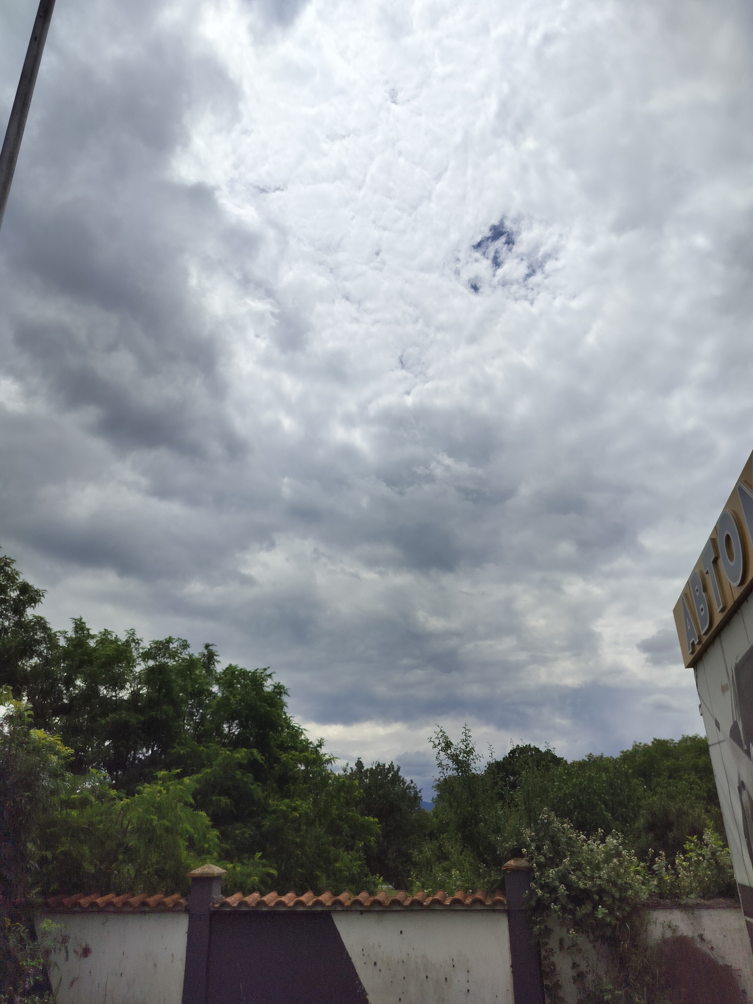 The image depicts a cloudy sky over a landscape in Tarnicheni, Bulgaria. In the foreground, a low, white wall with a red-tiled roof runs horizontally across the frame. Behind the wall, dense green trees form a thick line. To the right of the image, a portion of a building with a yellow sign displaying the word "ABTO" (likely a fragment of "Автосервиз", meaning "car service" in Bulgarian) is visible. The sky is filled with a mix of dark and light grey clouds, with a small patch of blue sky visible in the upper center. A vertical pole is present on the far left side of the image. The overall atmosphere suggests overcast or potentially rainy weather. There are no people or animals visible in the scene.