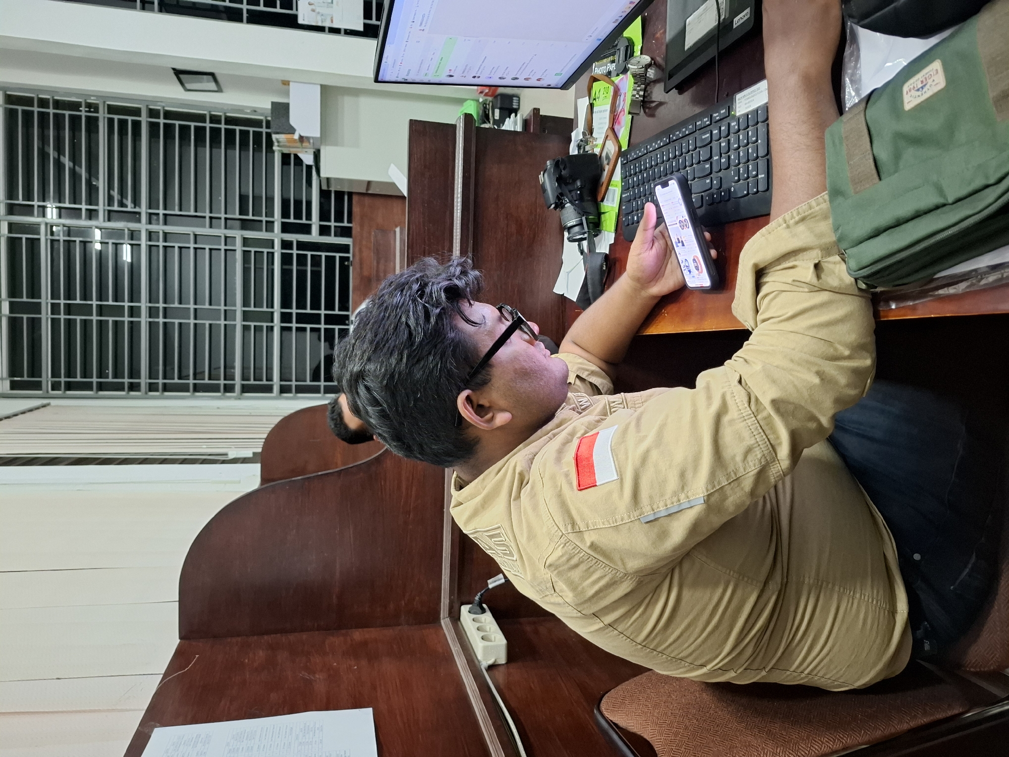 The image captures a person seated at a desk in what appears to be an office setting. The individual is holding a smartphone in their hands, gazing at the screen, while simultaneously positioned near a computer monitor. A keyboard and camera are situated on the desk, along with a green bag. The person wears a khaki-colored jacket with a rectangular emblem near the shoulder. The scene takes place within a cubicle. The presence of an Indonesian flag patch on the jacket, combined with the request for location, hints at the possibility that this photo was taken in Lhokseumawe, Indonesia. A window with a barred exterior is visible in the background.